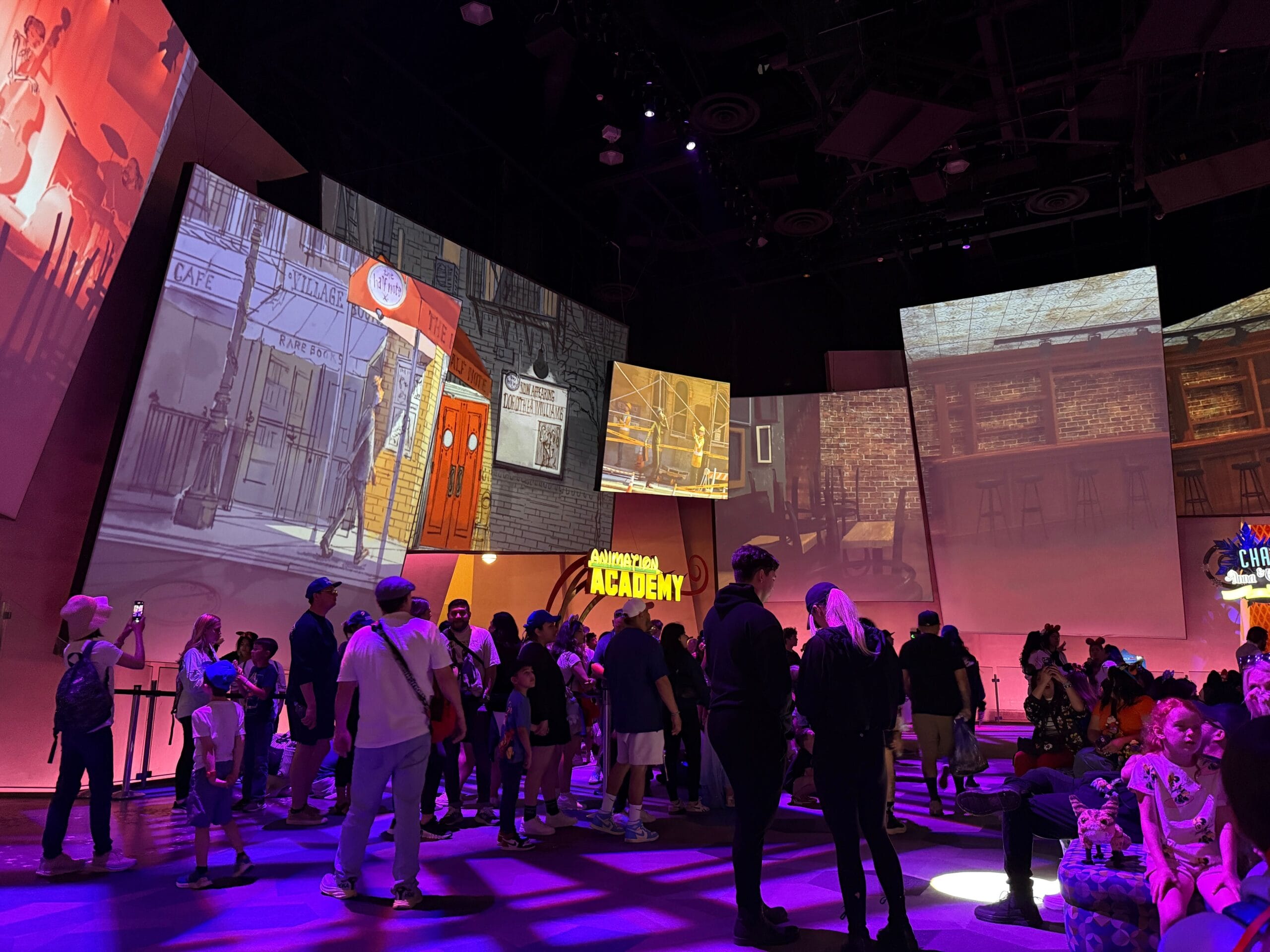 A crowd gathers inside a brightly lit room with animated cityscapes projected on large screens above.