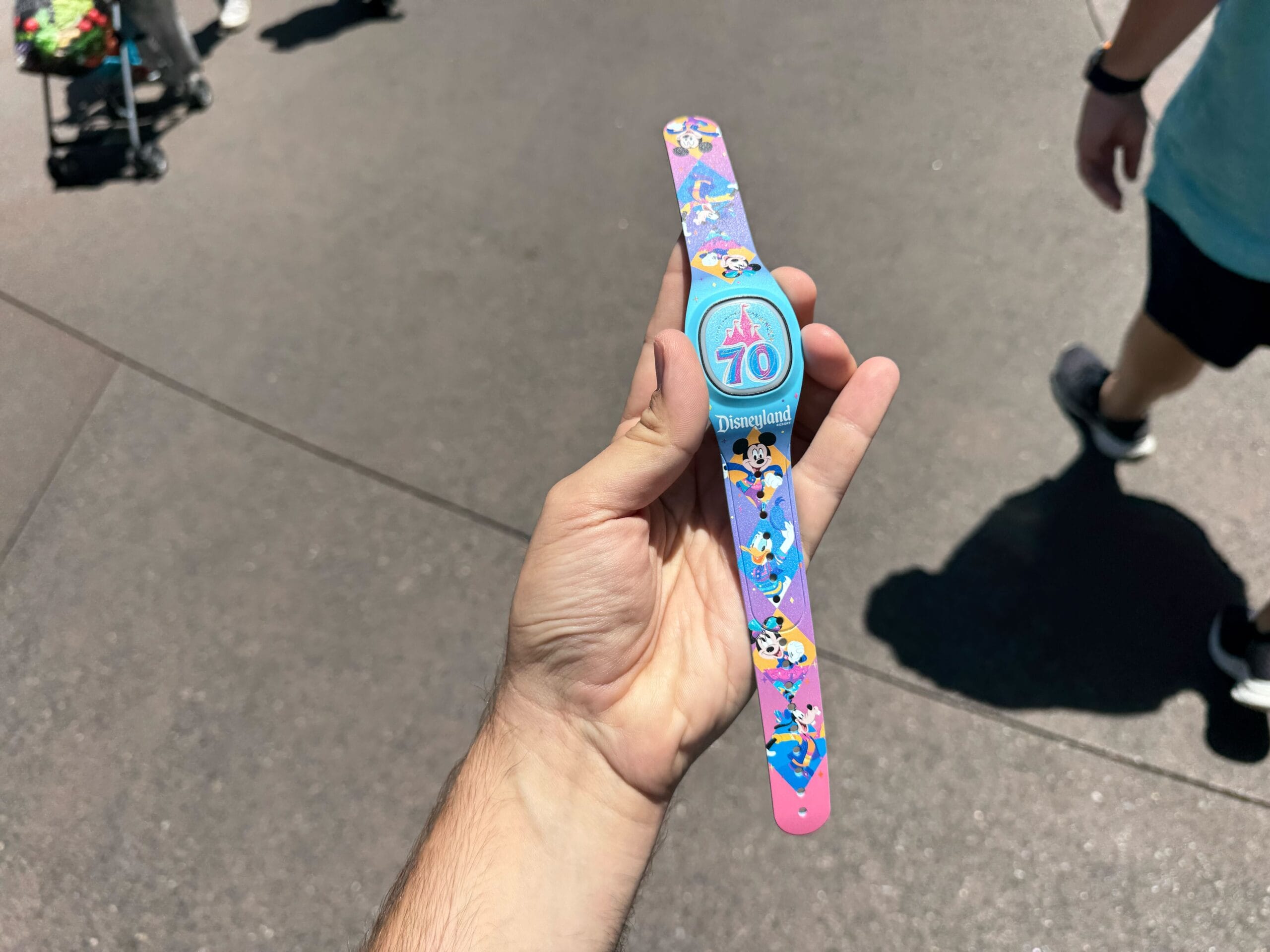 Hand holding a colorful Disneyland Resort 70th Anniversary MagicBand+ with Disney characters, pavement in background.