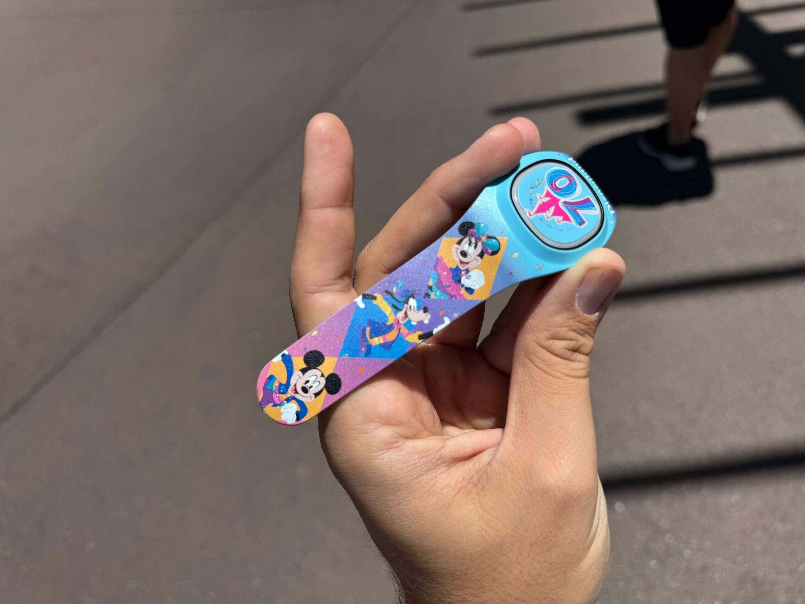 A hand holds a MagicBand+ featuring Mickey, Minnie, and Donald on a colorful Disneyland Resort background.