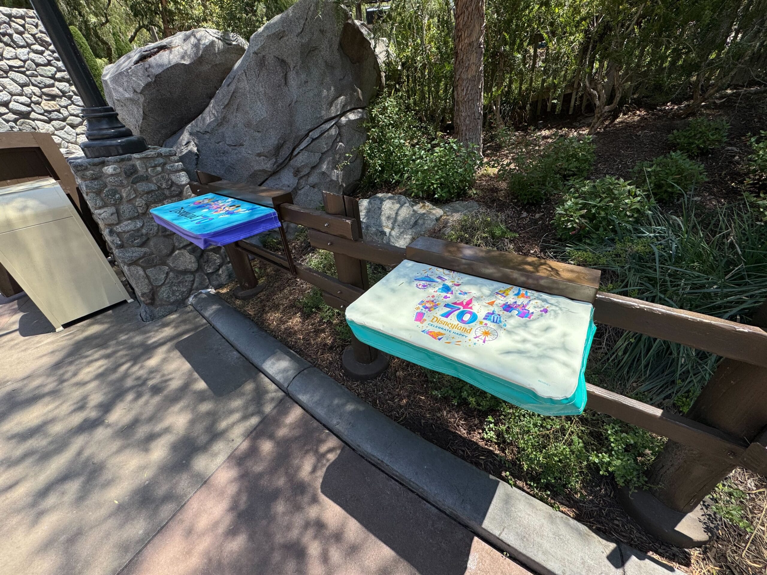 Two tables with colorful covers, one reading "70," beside a wooden fence in a shaded outdoor area.