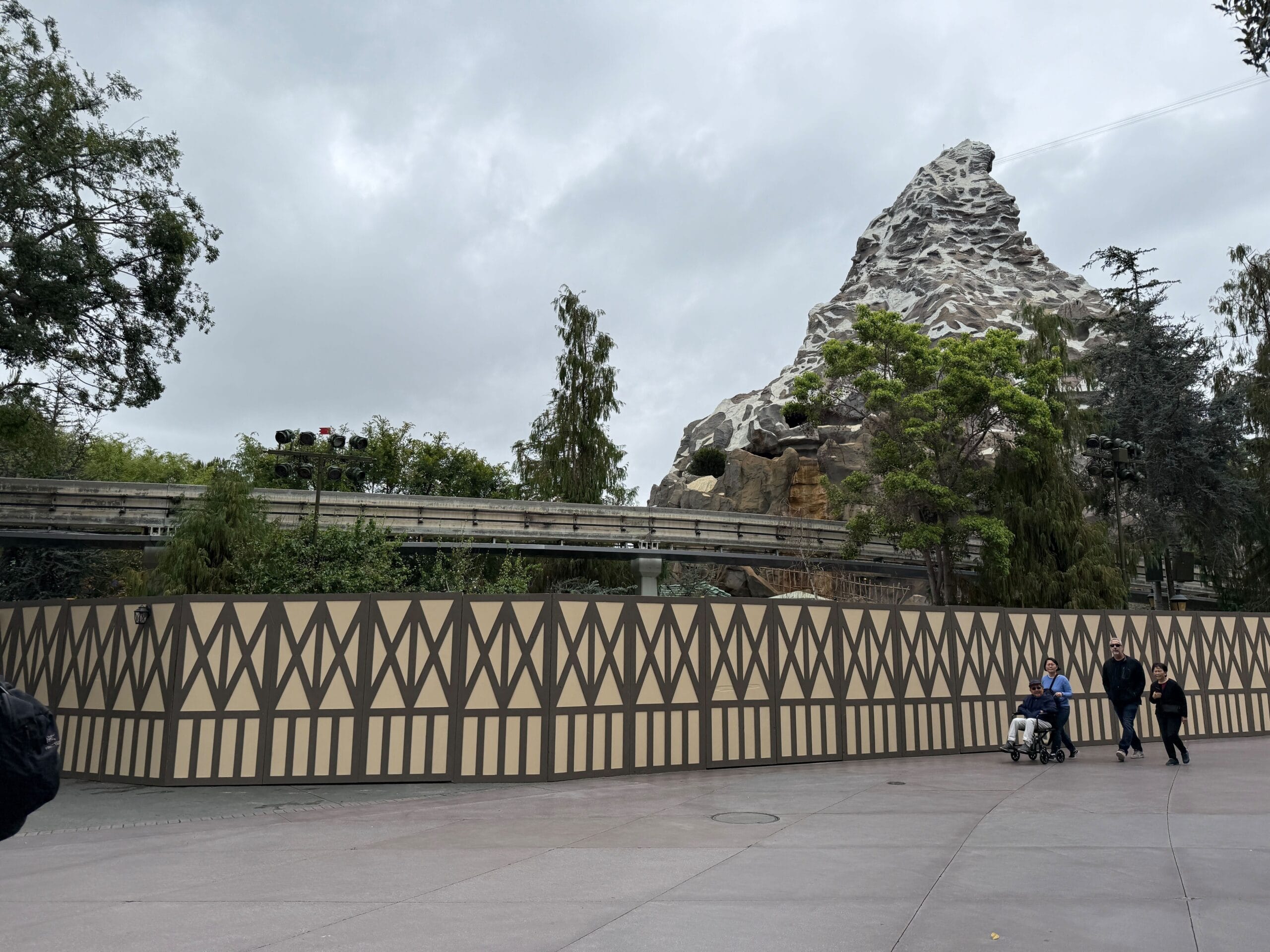 A construction wall blocks part of a mountain-themed roller coaster ride on a cloudy day, with a few people walking nearby.