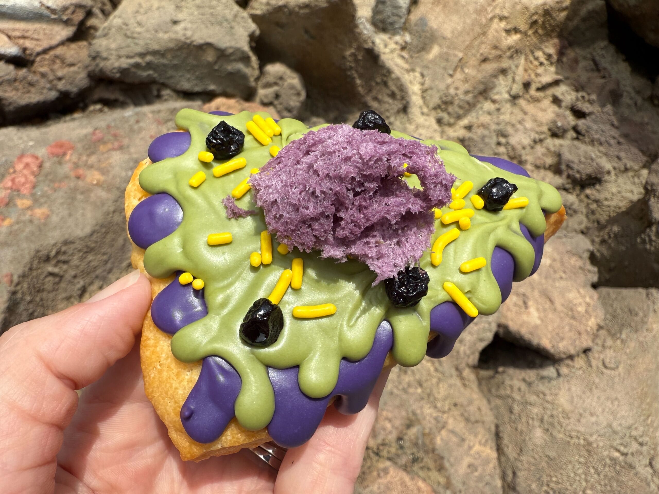 A hand holds a triangular pastry topped with purple, green, and yellow icing, sprinkles, and a purple filling.