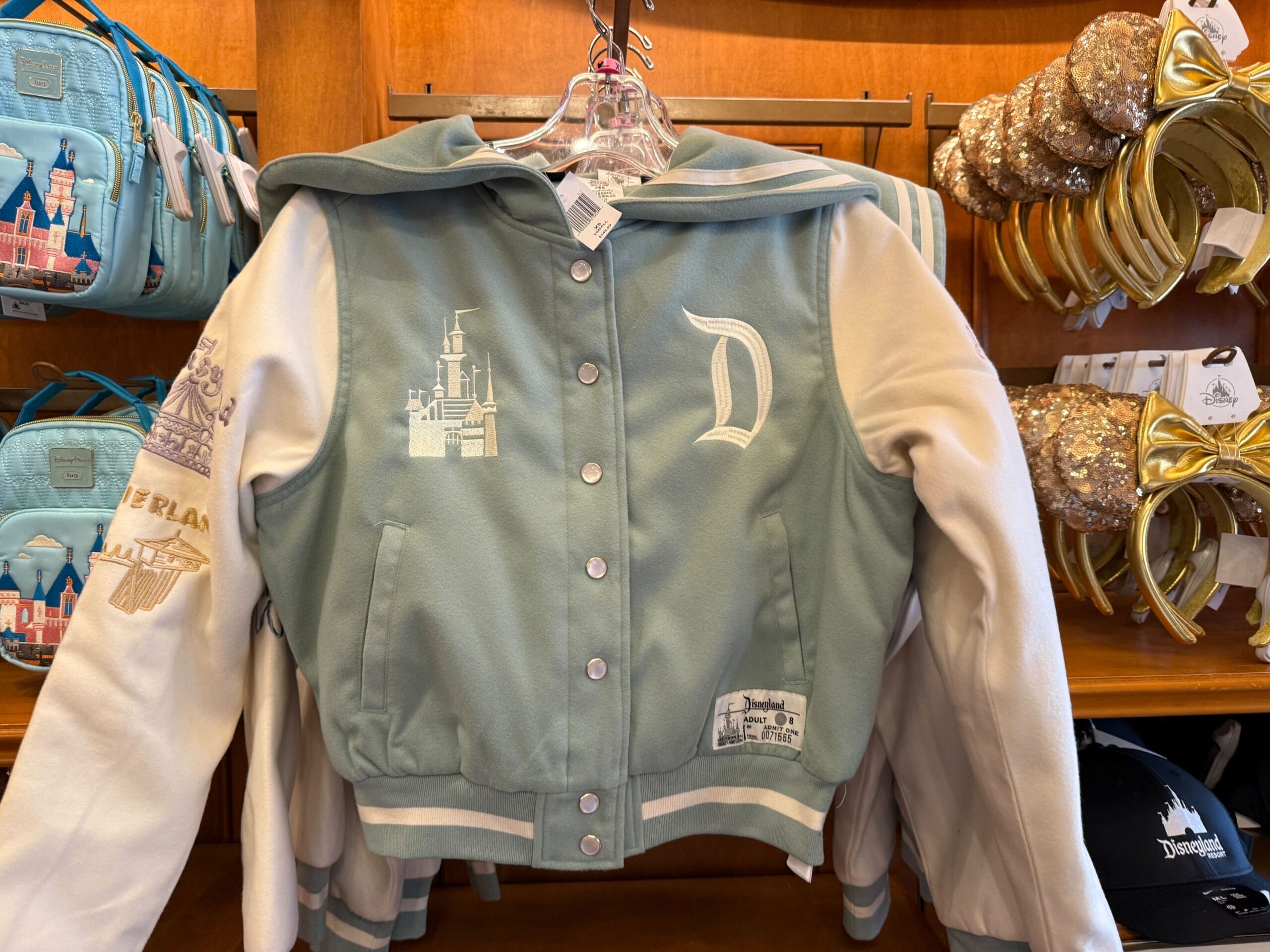 A pastel green and white Disneyland jacket with castle and "D" patches, hanging on a store rack.