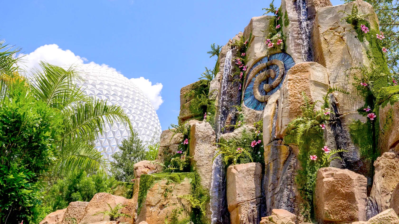 Rocky waterfall with pink flowers, greenery, and Moana emblem at EPCOT