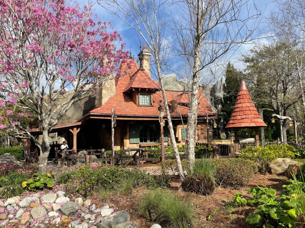 A storybook-style cottage with a red roof, surrounded by blooming trees and lush garden greenery. A storybook-style cottage with a red roof, surrounded by blooming trees and lush garden greenery in Fantasyland.