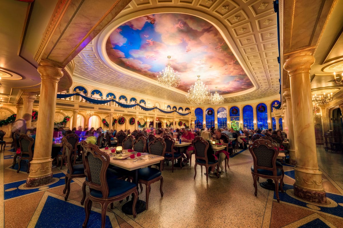 Elegant restaurant with ornate chandeliers, painted ceiling, and diners seated at tables under warm lighting. Fantasyland Be our Guest restaurant with ornate chandeliers, painted ceiling, and diners seated at tables under warm lighting.