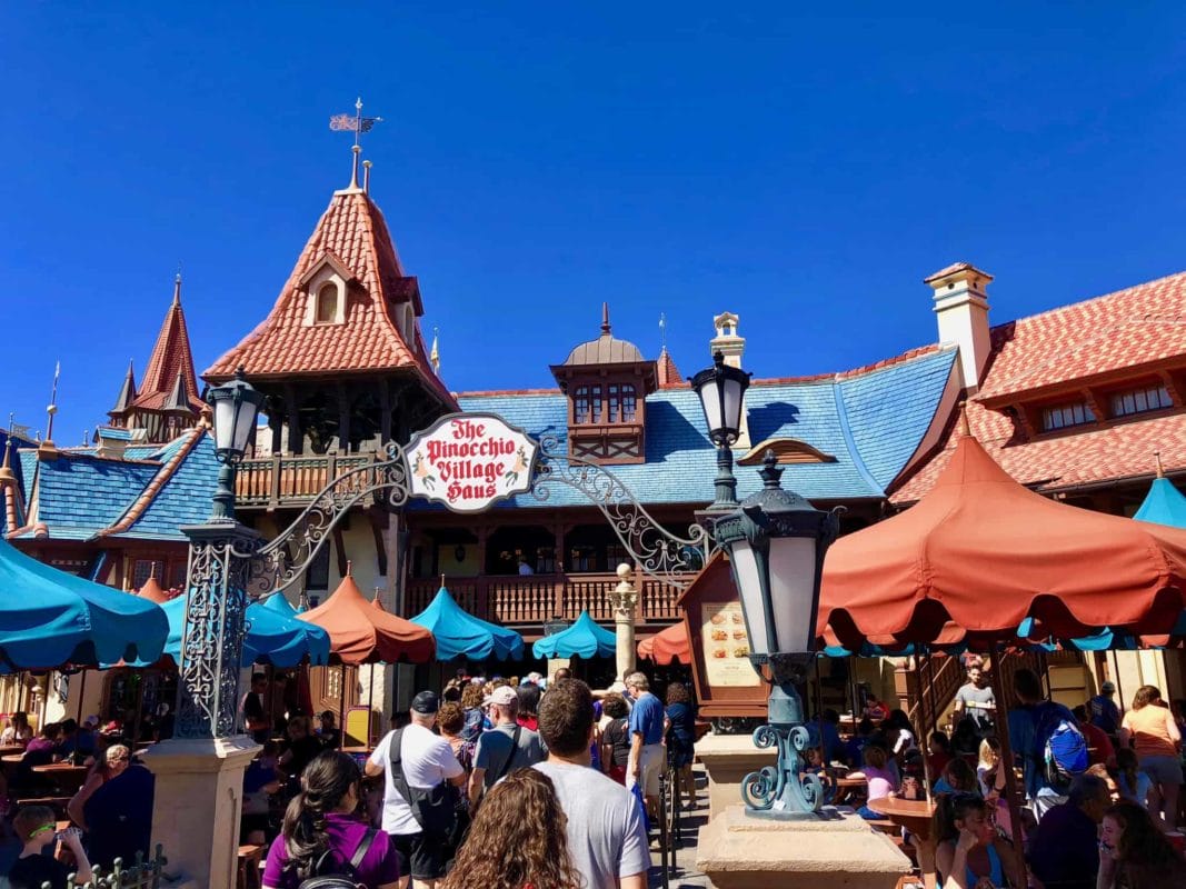 Crowds gather outside The Pinocchio Village Haus restaurant with colorful umbrellas and a fairytale-style building under blue sky. Crowds gather outside The Pinocchio Village Haus restaurant in Fantasyland with colorful umbrellas and a fairytale-style building under blue sky.