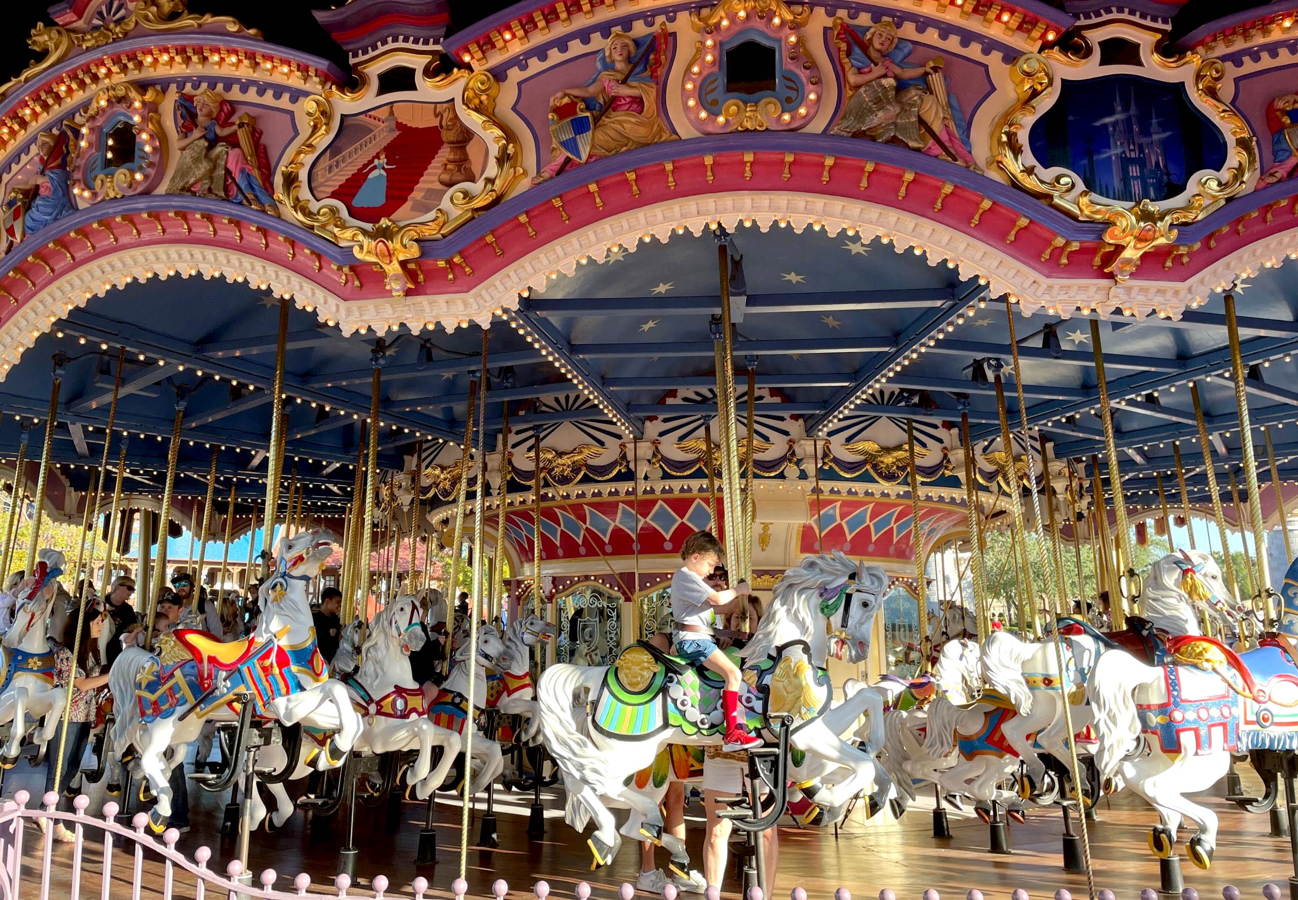 Children ride white carousel horses on a colorful, ornate merry-go-round under bright sunlight at an amusement park. Prince Charming Regal Carrousel in Fantasyland at the magic kingdom.