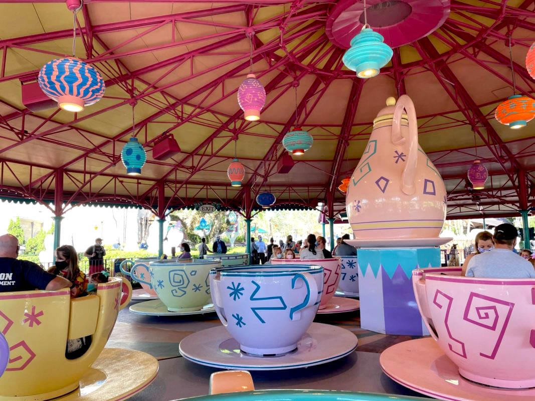 People sit in large, colorful teacups under a whimsical, lantern-lit canopy at an amusement park ride. People sit in large, colorful teacups under a whimsical, lantern-lit canopy in Fantasyland
