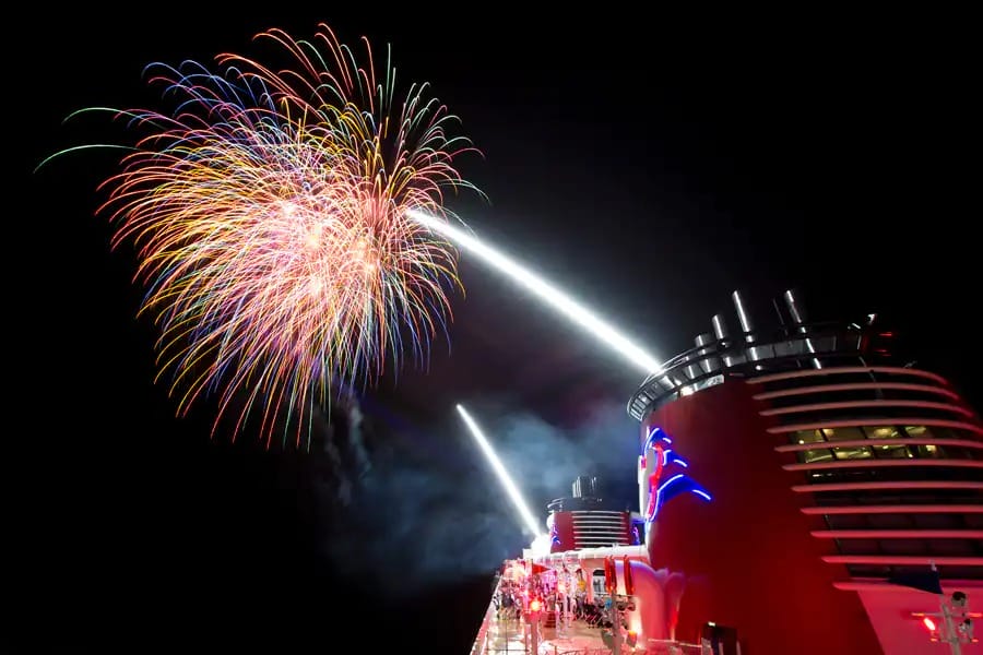 Colorful fireworks burst over a Disney Cruise deck at night, with bright spotlights beaming into the sky.