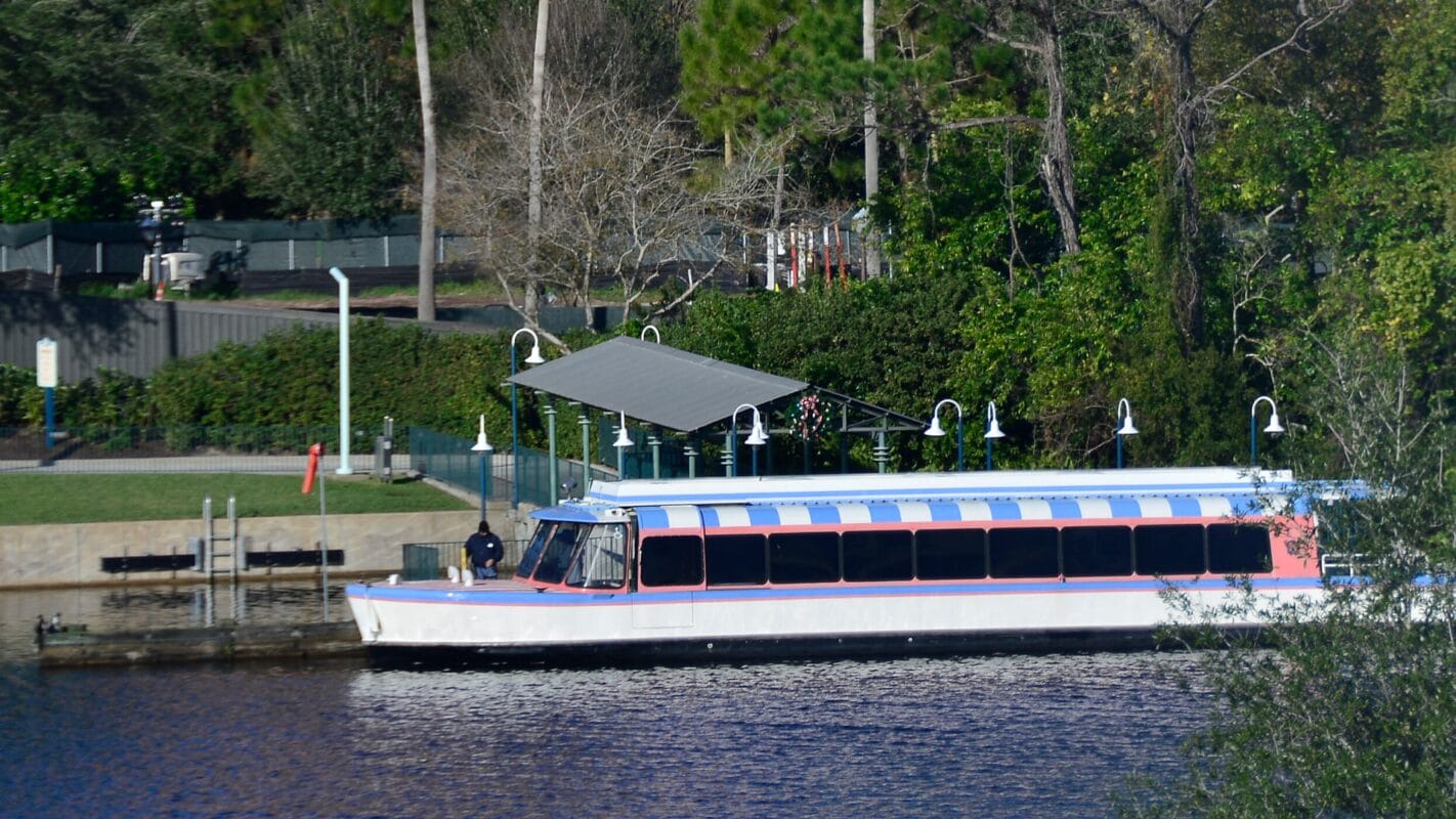A boat with blue and red stripes is docked near trees and the Hollywood Studios entrance