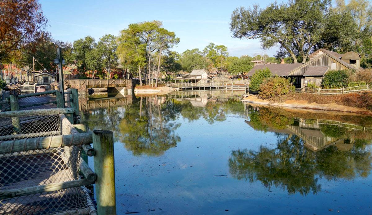 A wooden bridge and rustic Frontierland buildings reflect in a calm river amid trees and greenery under a clear sky.