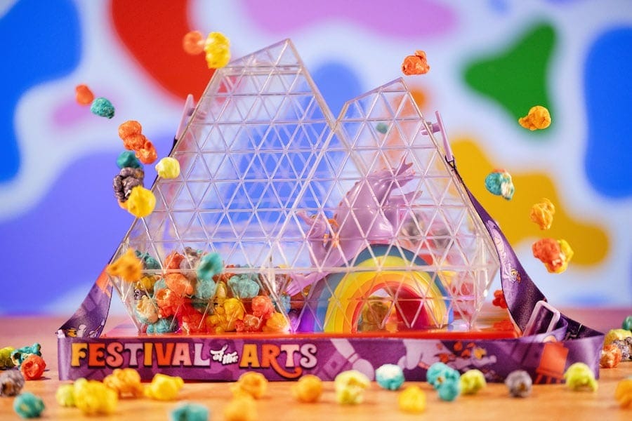 Colorful popcorn spills from the Imagination Pavilion Disney Popcorn Bucket, with a rainbow and "Festival of the Arts" lanyard in front.