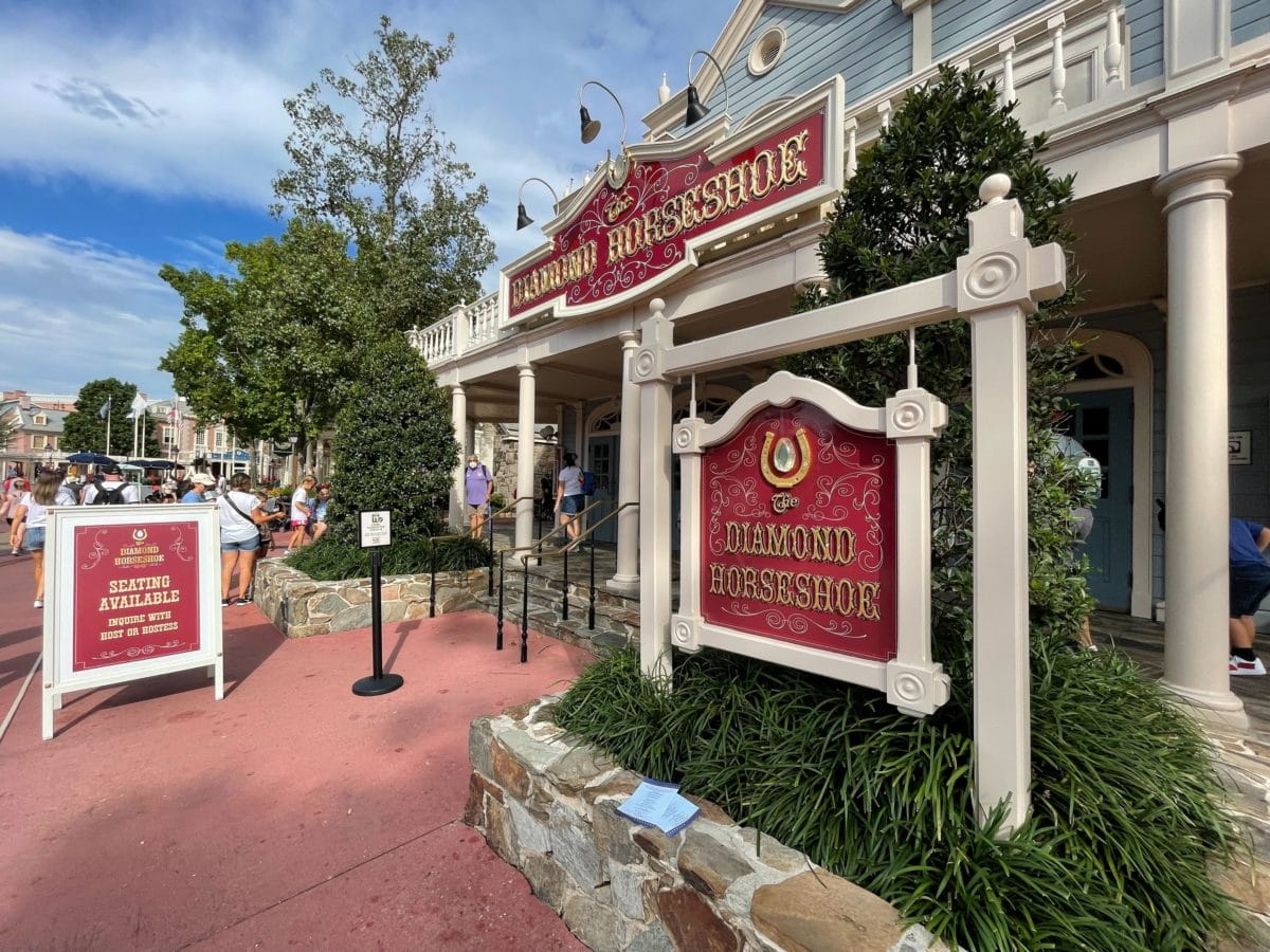 Entrance of the Diamond Horseshoe restaurant in Liberty Square, with outdoor signs and people strolling by on a sunny day.