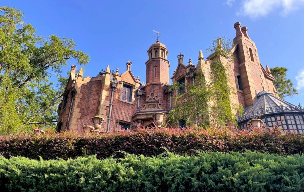 A stately, brick mansion with gothic architecture evokes Magic Kingdom charm amid lush landscaping and a clear blue sky.