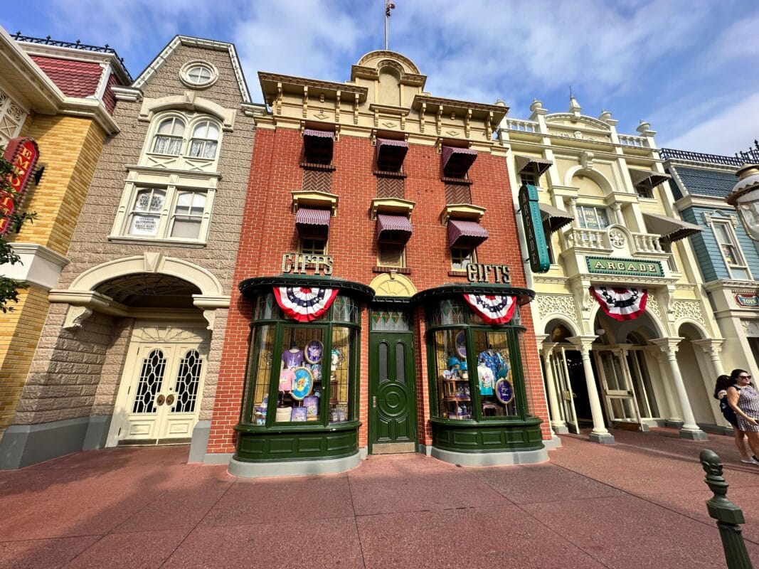 A brick gift shop on Main Street USA, adorned with patriotic bunting, sits among ornate buildings under a blue sky.