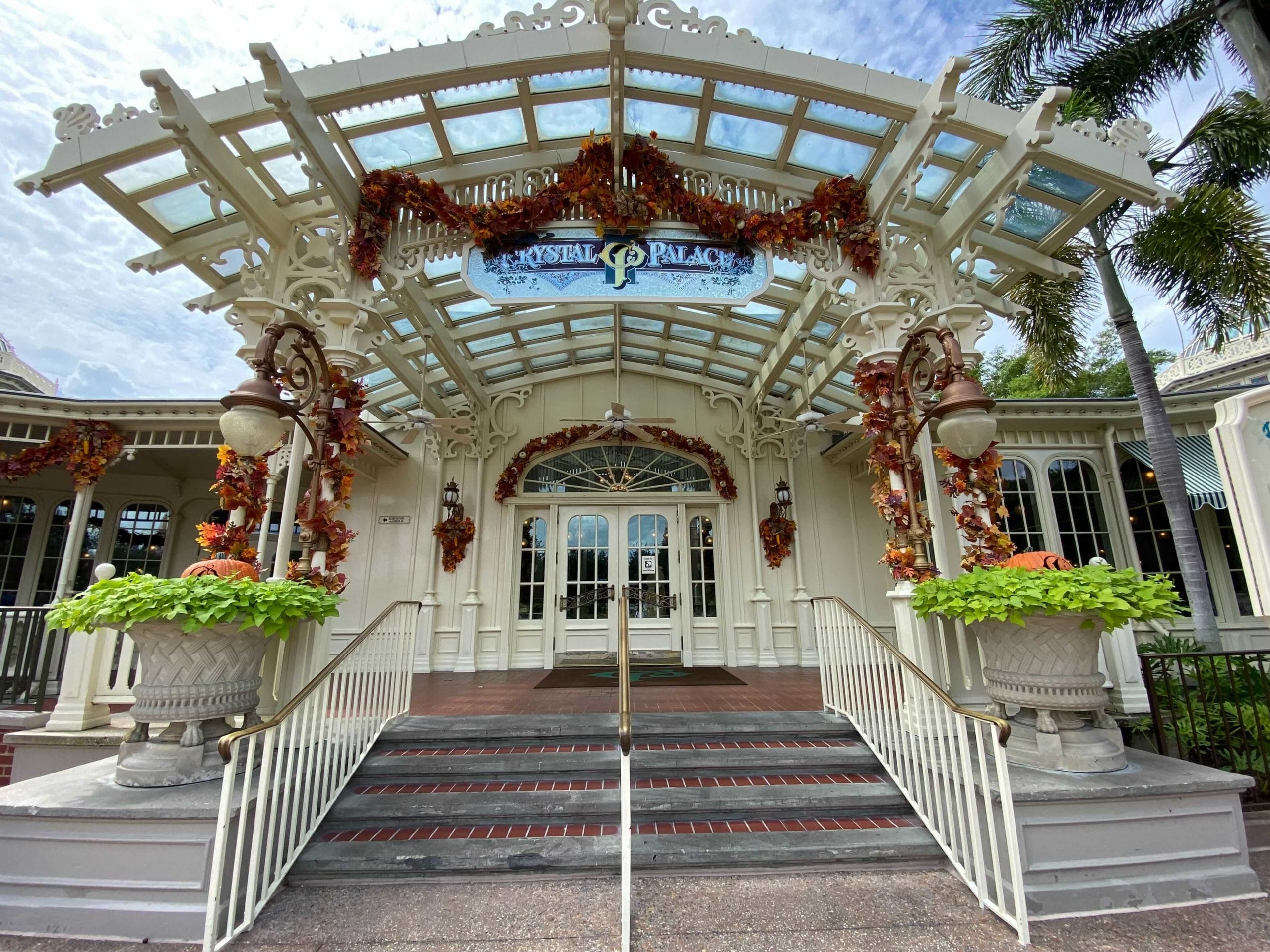 Ornate entrance of the Crystal Palace on Main Street USA, adorned with autumn leaves and plants under a glass canopy.