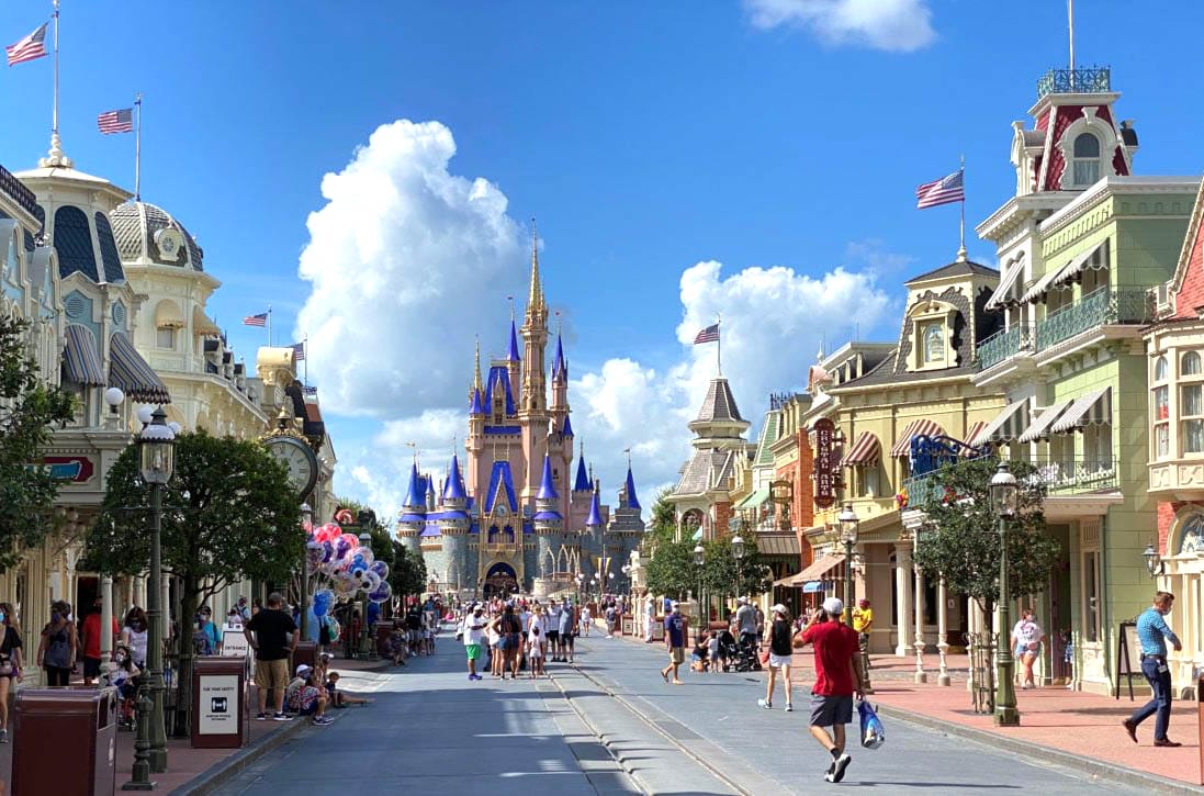 People stroll down Main Street USA toward Cinderella Castle at Magic Kingdom, with shops and balloons lining the sunny walkway.