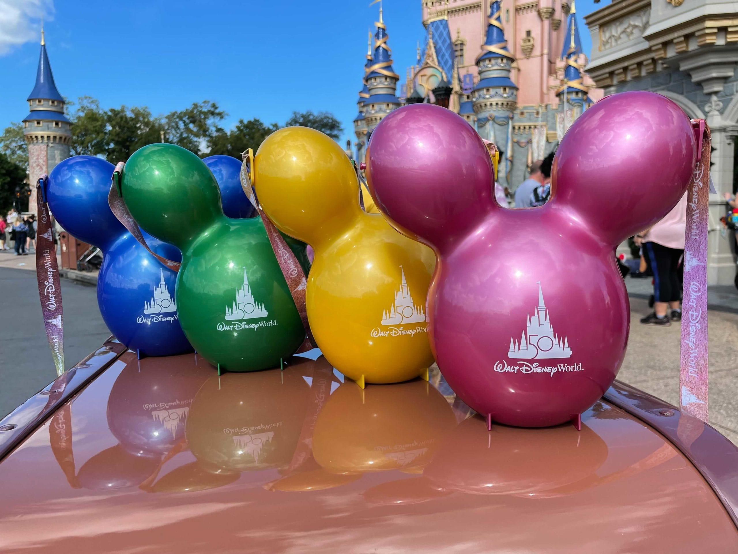 Four colorful Mickey Mouse balloons with “50th Walt Disney World” float before Cinderella Castle, beside a Disney Popcorn Bucket.