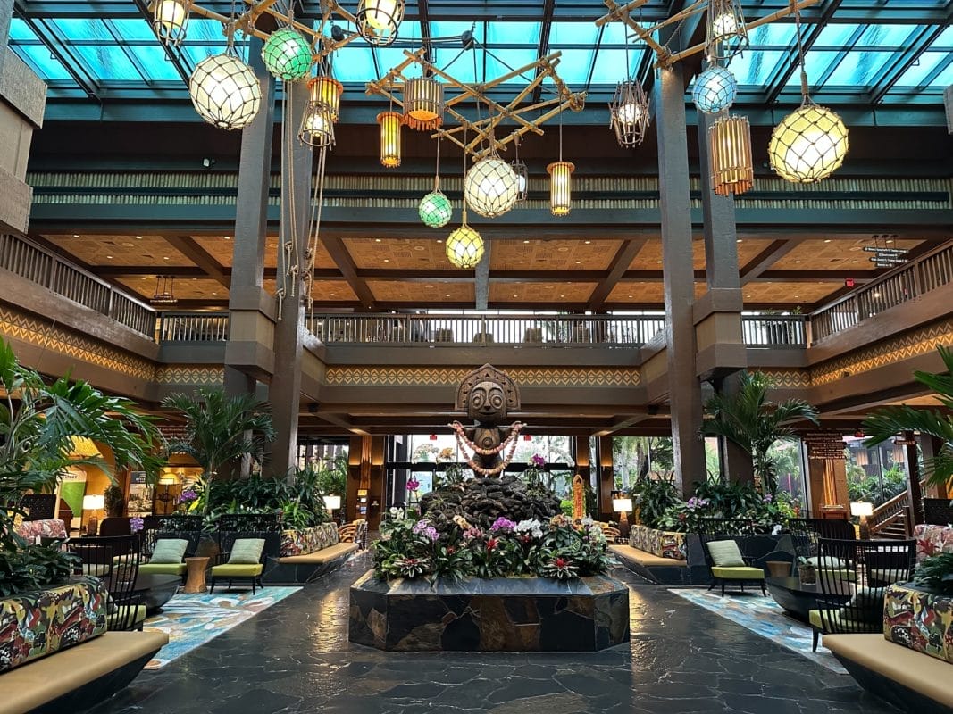 Spacious disney deluxe resort lobby with tropical plants, tiki statue centerpiece, and lanterns under a glass ceiling.