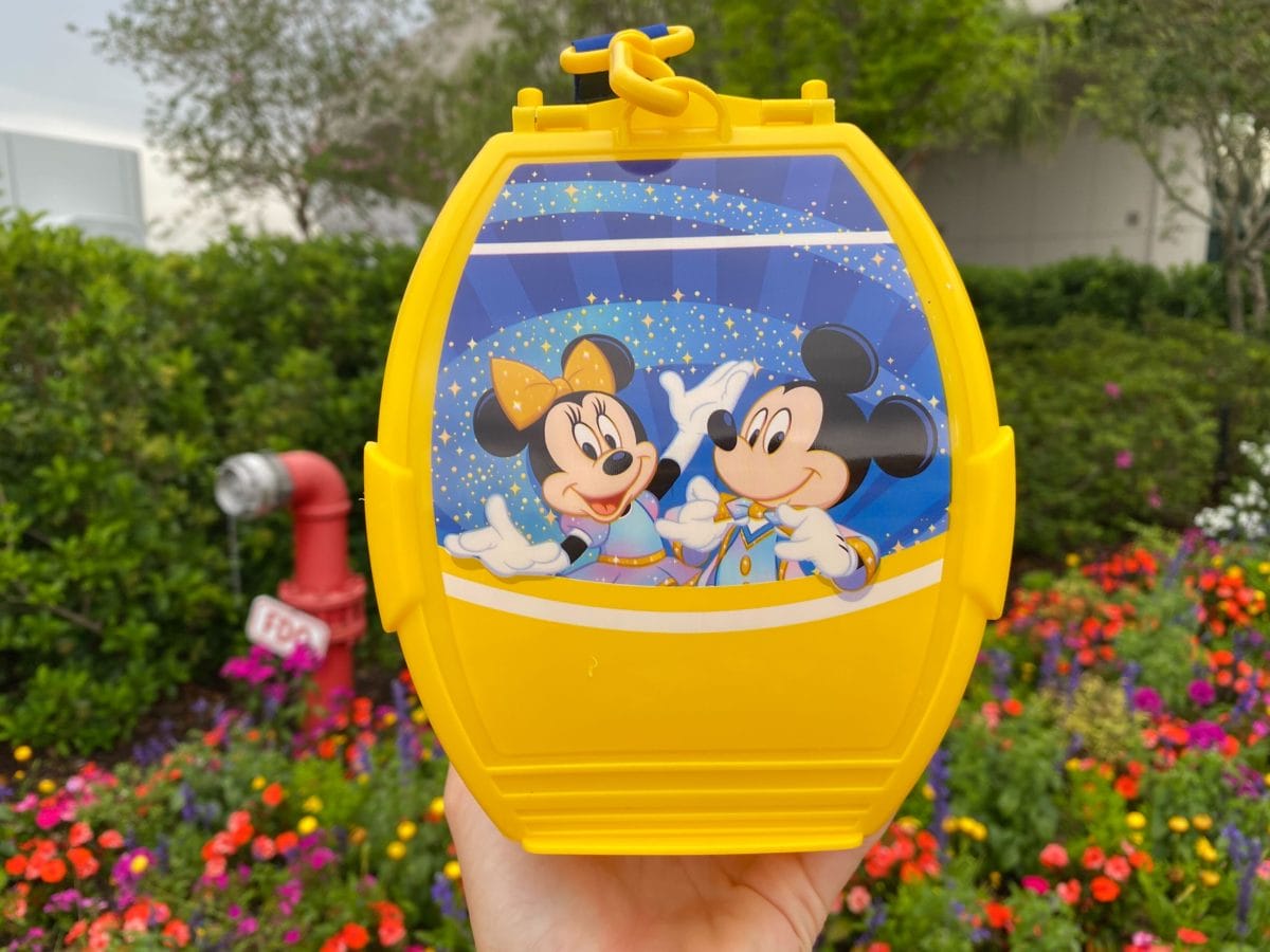 A Disney Popcorn Bucket featuring Mickey and Minnie Mouse, held before vibrant flowers and lush greenery.