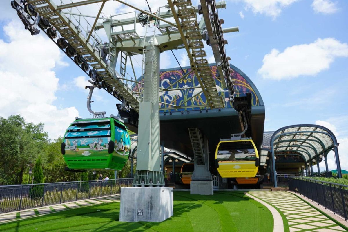 Colorful Disney Skyliner gondolas arrive at a decorative station under a bright, partly cloudy sky at the Epcot station.