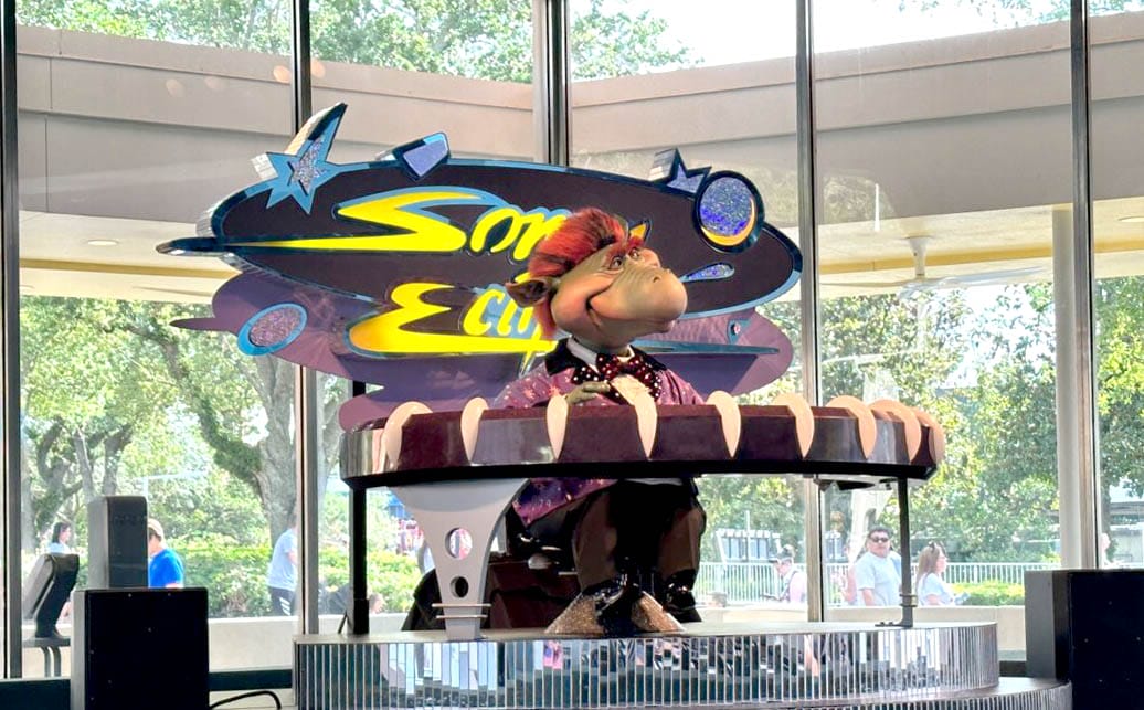 A cartoon dinosaur animatronic performs on a futuristic Tomorrowland stage before a colorful "Sonny Eclipse" sign. Sonny Eclipse performs on a futuristic Tomorrowland stage