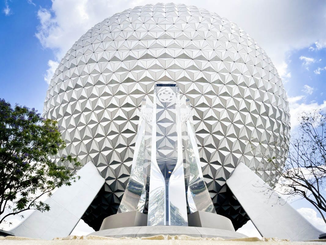 A large geodesic sphere at EPCOT with reflective sculpture and trees in the foreground under a partly cloudy sky.