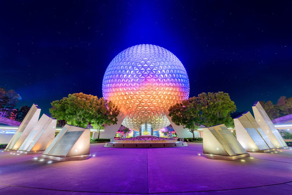 Spaceship Earth at Epcot, Disney World, shines with colorful lights at night, framed by trees and monuments.