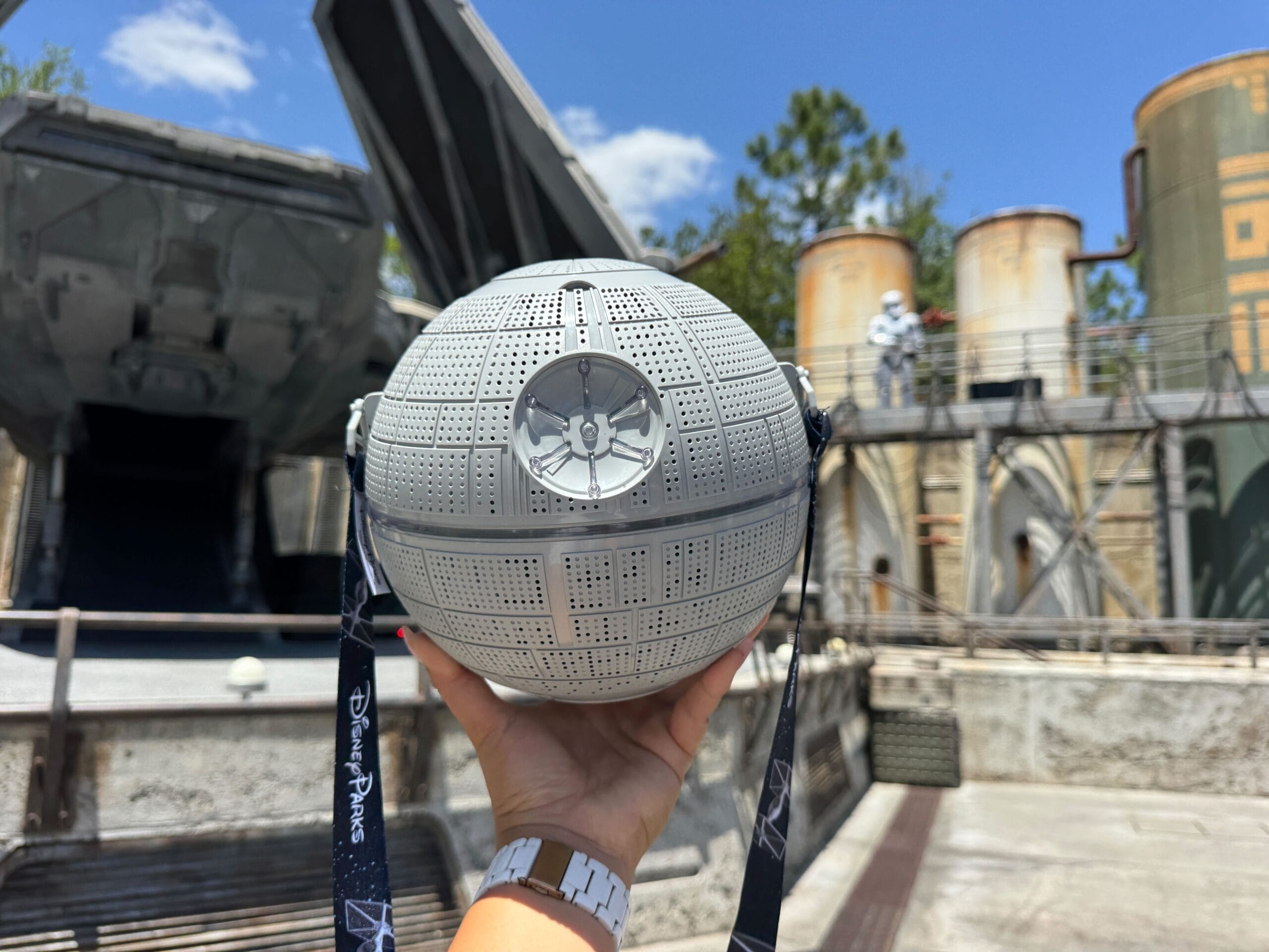 A hand holds a Disney Popcorn Bucket shaped like the Death Star at a Star Wars-themed park under a blue sky.