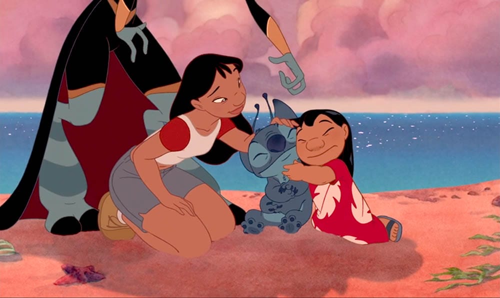 Two girls and a blue alien hug and smile on a sandy beach—perfect Stitch wallpaper for free downloads.