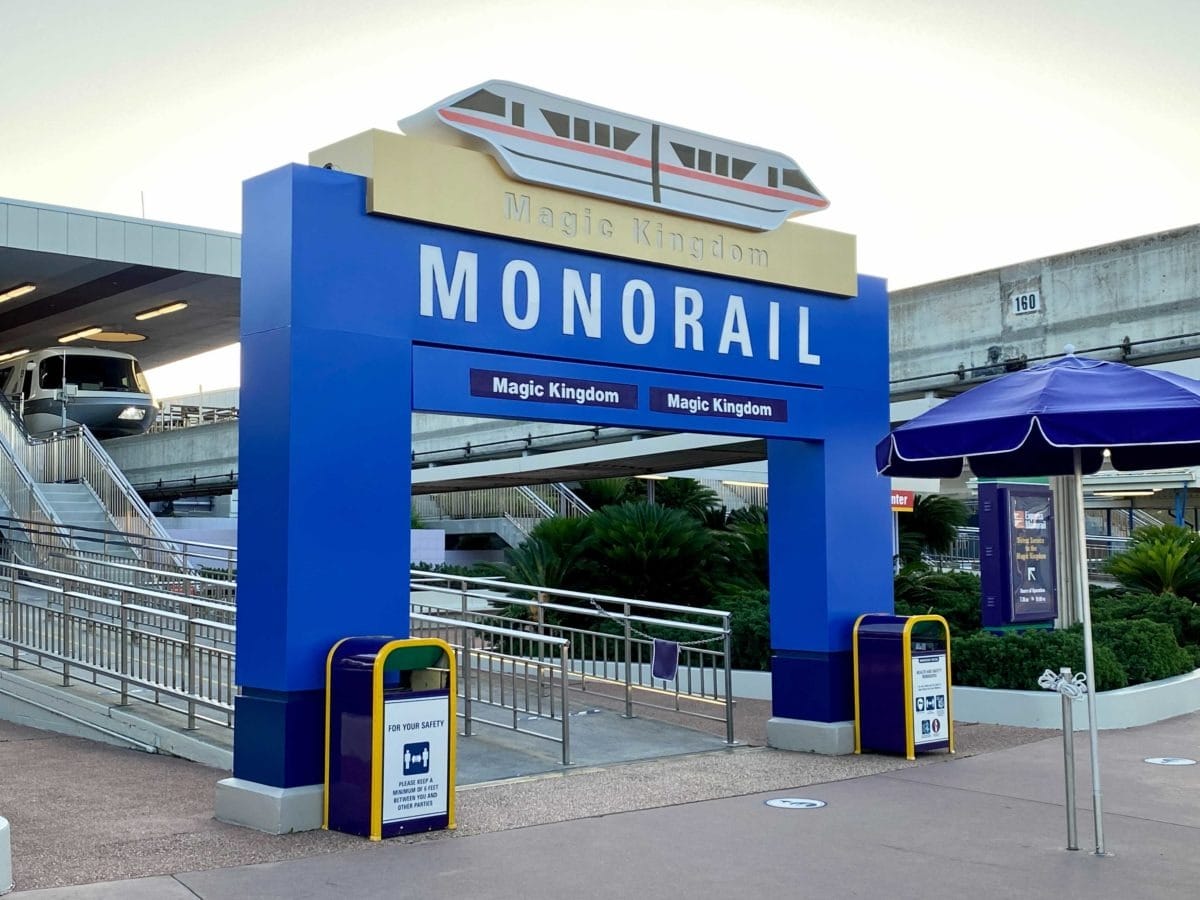 Blue Magic Kingdom Monorail entrance sign with Disney monorail train on left and purple umbrella on right at ttc past magic kingdom toll plaza