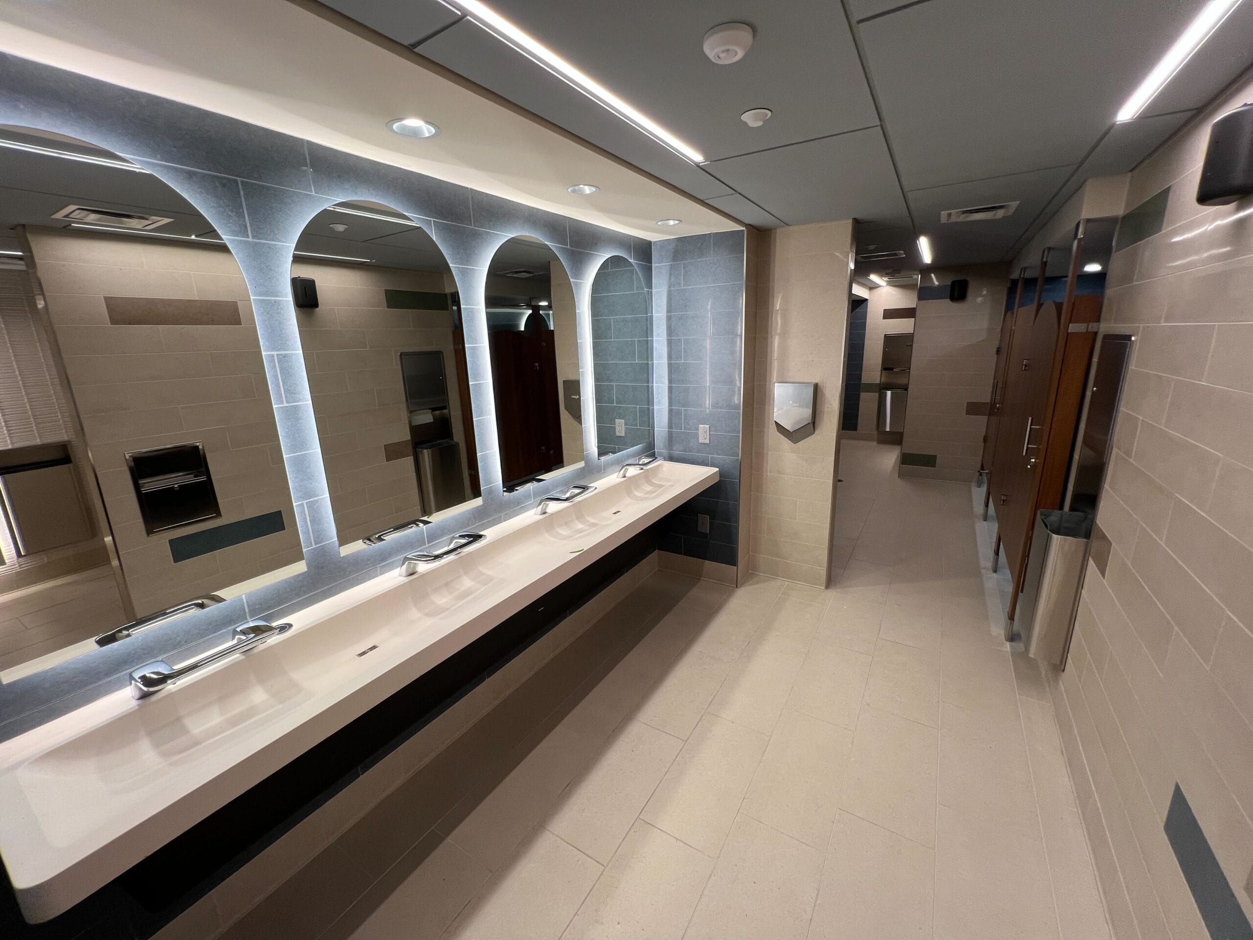 Refurbished public restroom with multiple sinks, mirrors, tiled walls, and several stalls in the background.