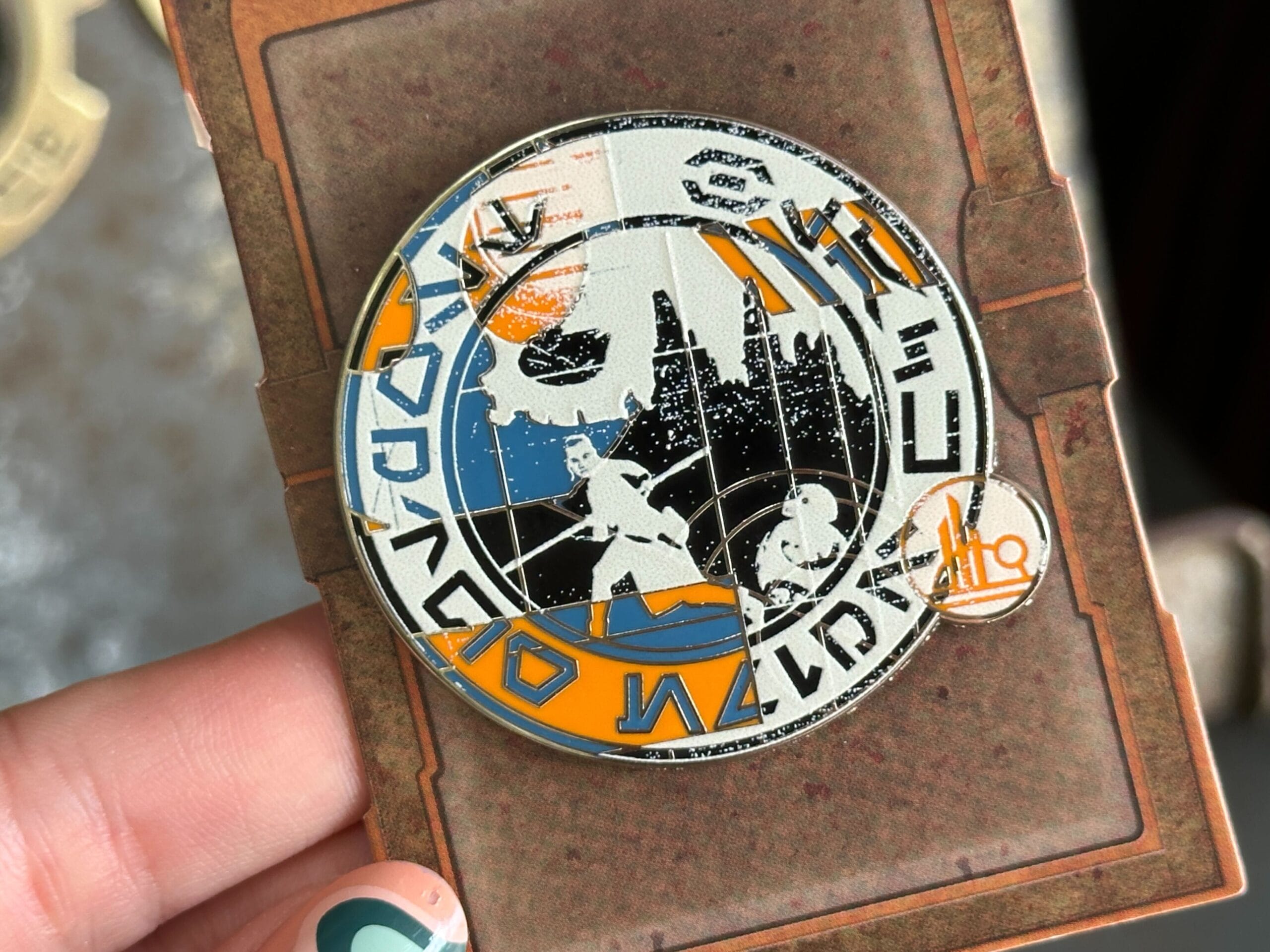 A hand holds a circular pin with Star Wars designs and Aurebesh text, featuring characters and spaceships.