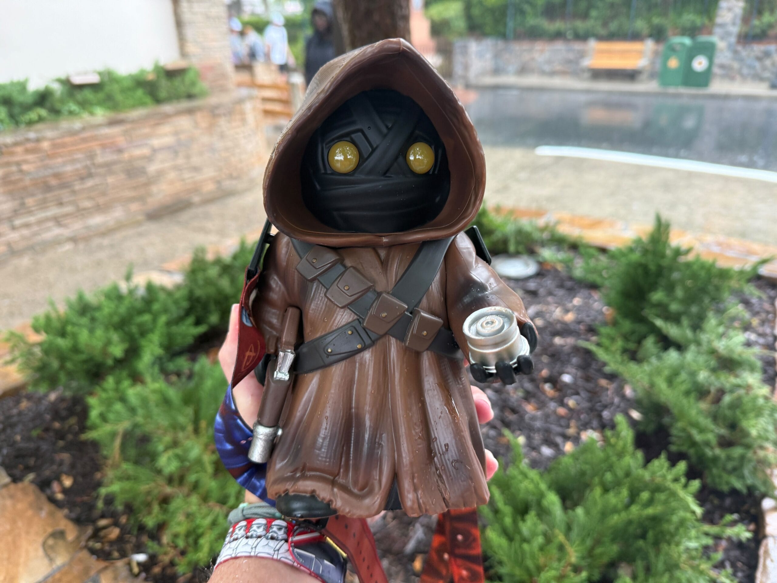 A hand holds a Jawa popcorn bucket from Star Wars, with greenery and a wet street in the background.