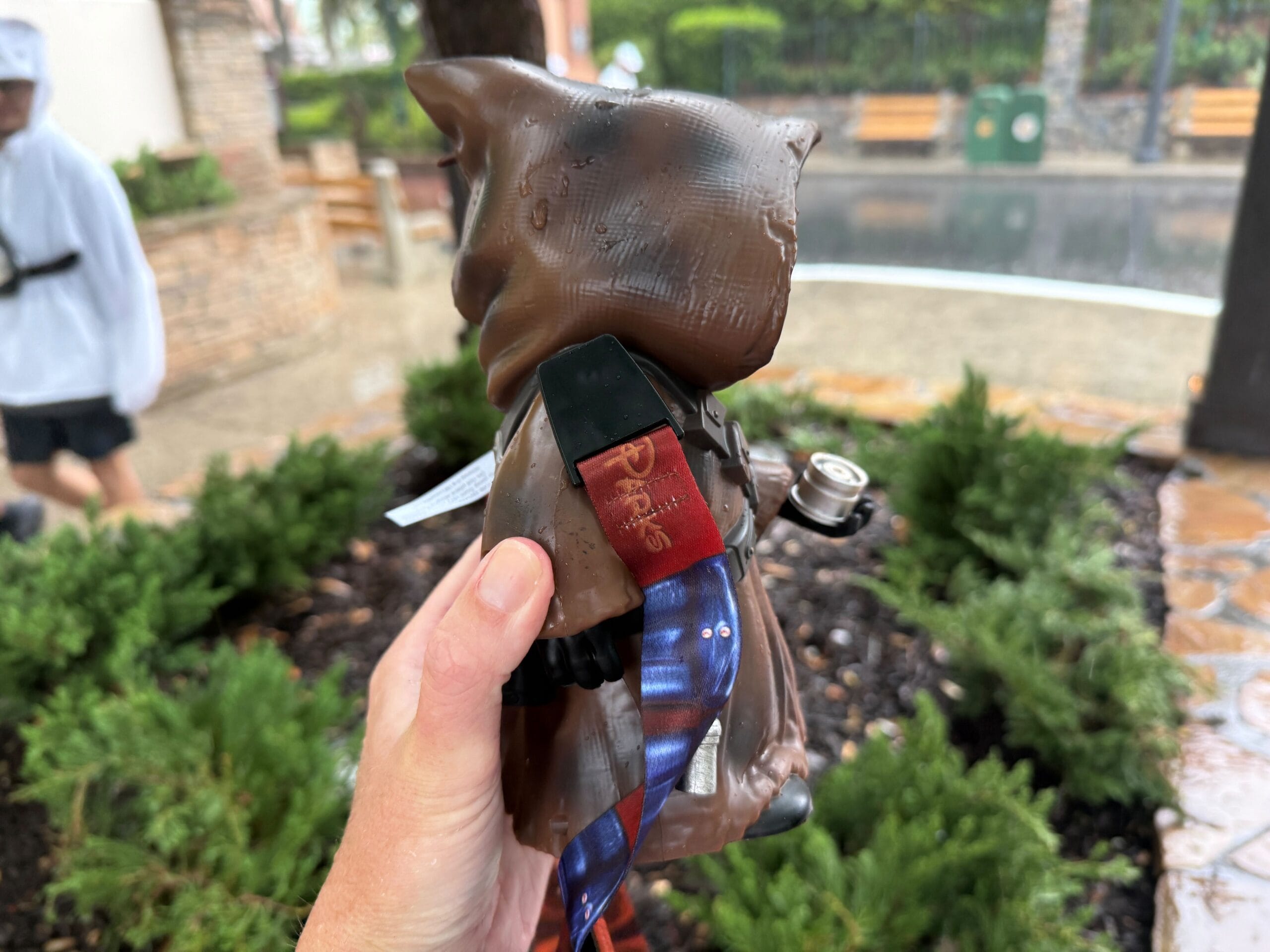 A hand holds a toy figure in a brown hooded robe outdoors on a rainy day, beside green bushes and stone paths.