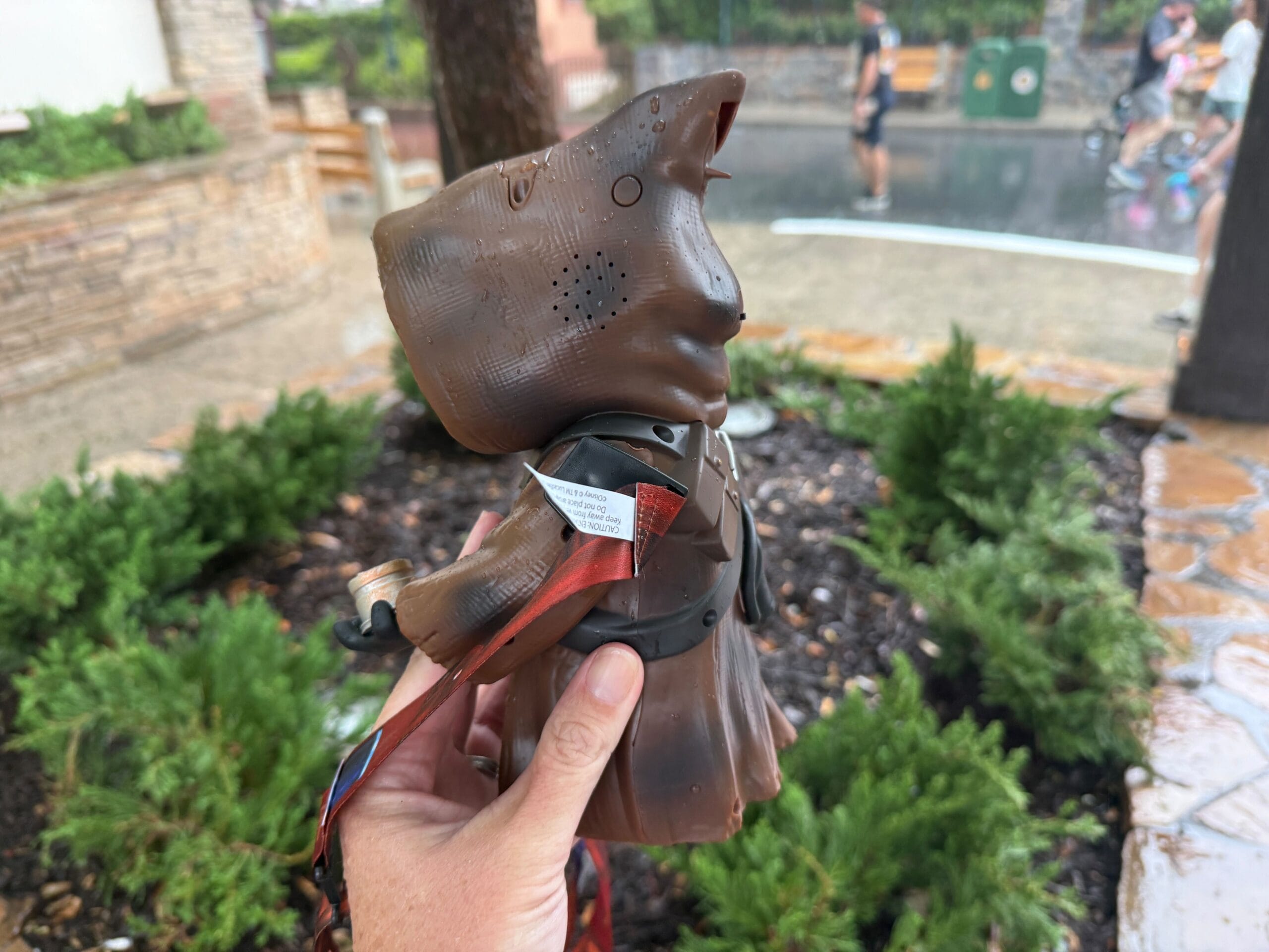 A hand holds a brown, robed Star Wars Jawa toy outdoors on a rainy day with wet ground and greenery.