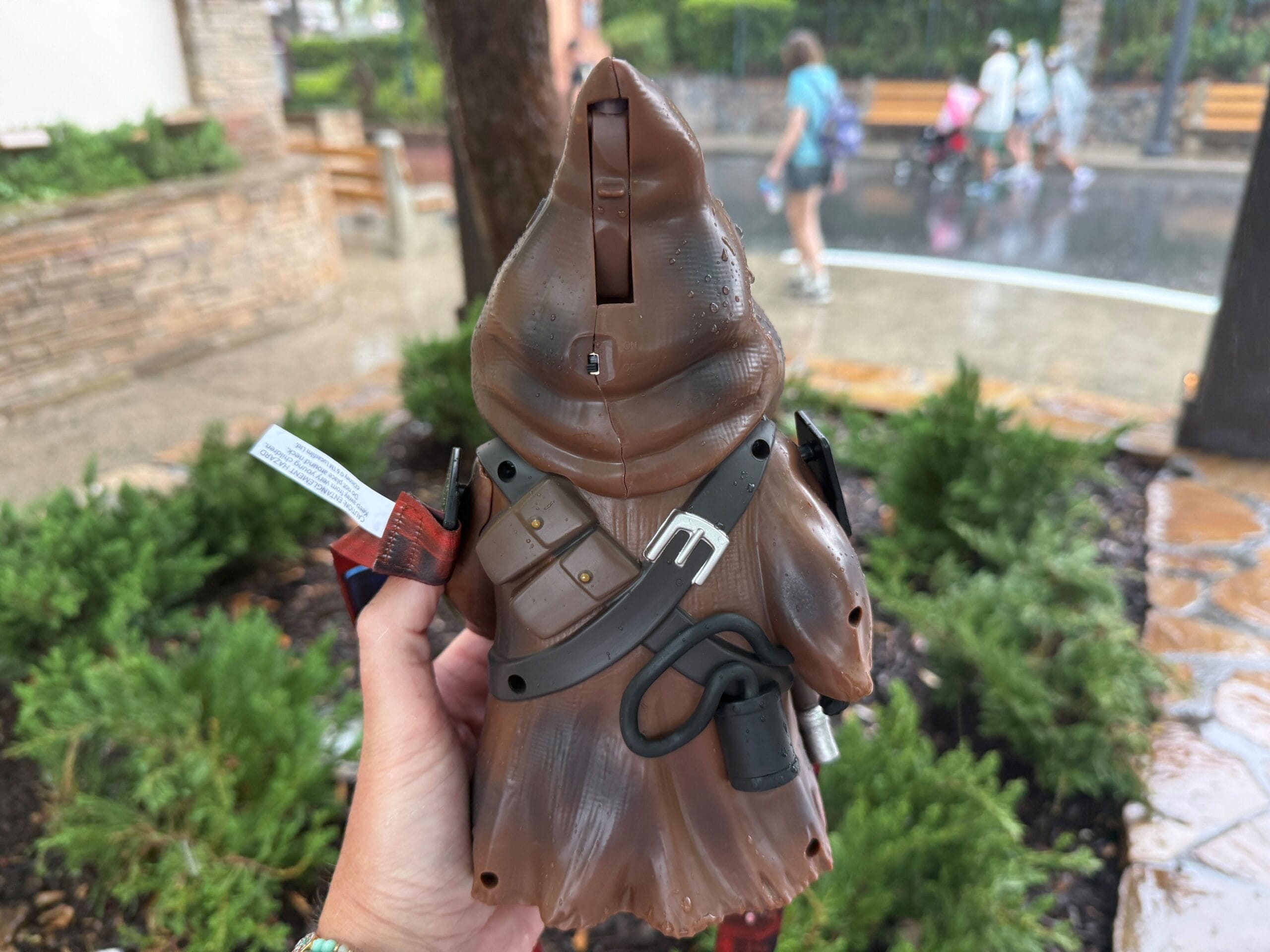 A hand holding a toy Jawa figure from Star Wars, with greenery and people in the background on a rainy day.