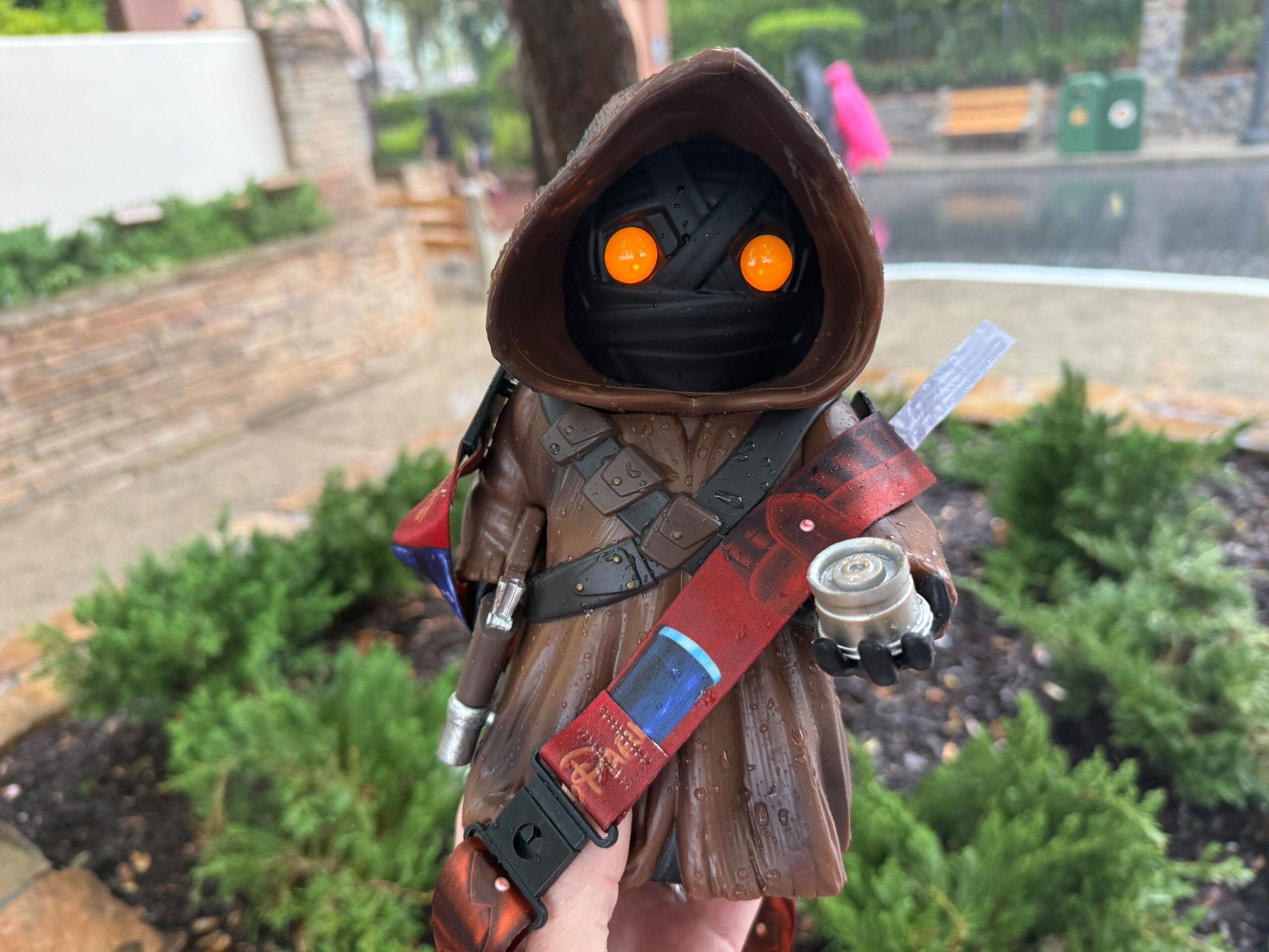 A hooded figure with glowing orange eyes and brown robes holding a small metal object outdoors on a rainy day.
