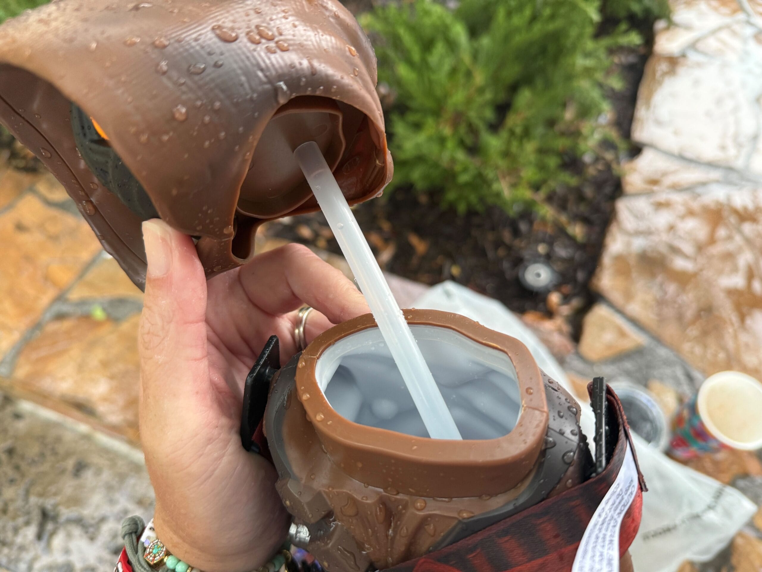 Hand holding an open brown water bottle with a straw, water droplets on the bottle, and greenery in the background.