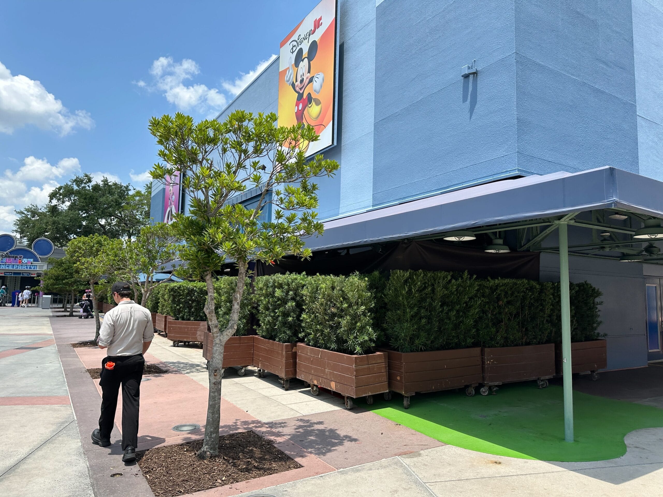 A man walks near planters and barriers outside a blue Disney building with a Mickey Mouse sign on a sunny day.