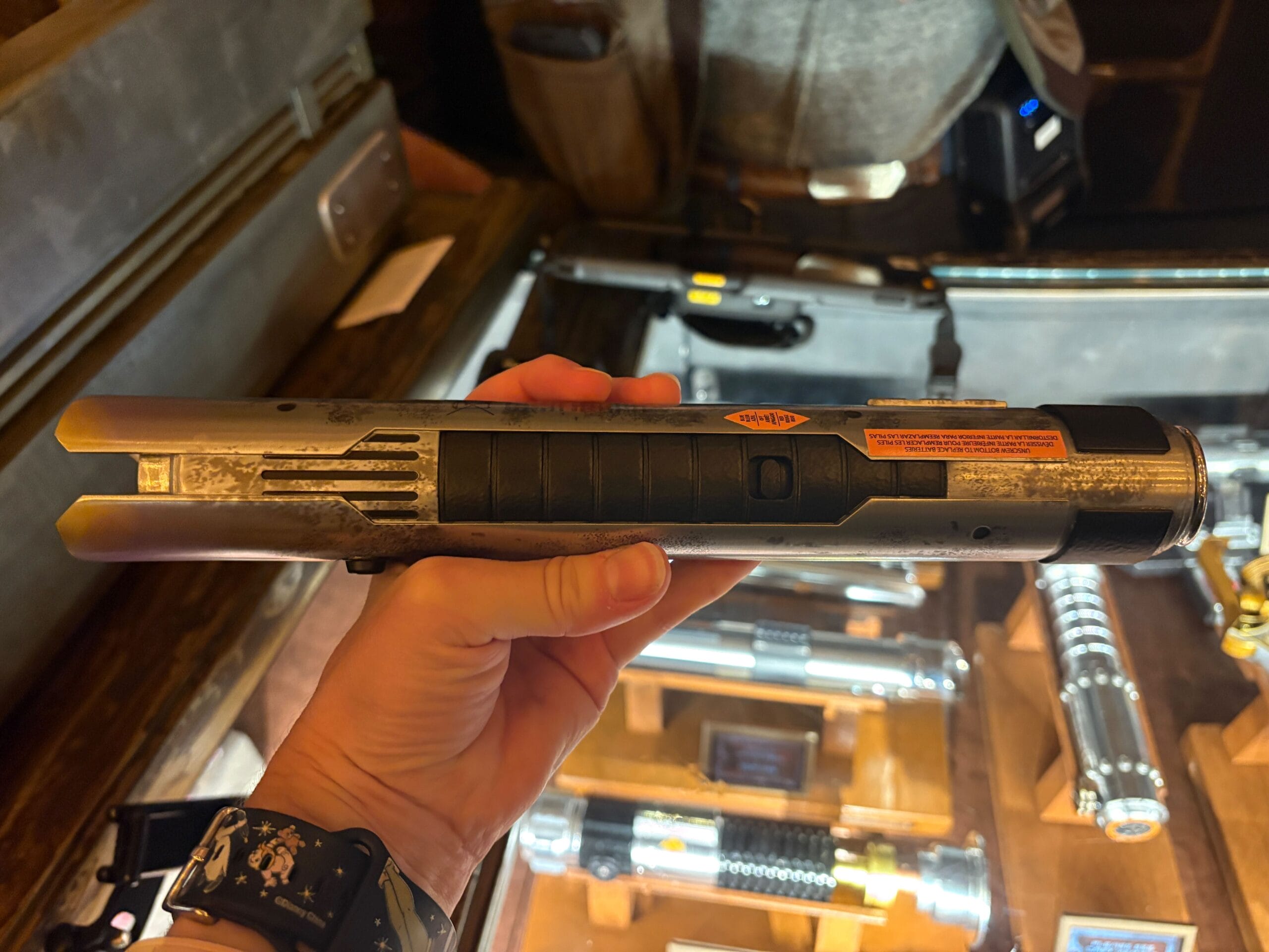 A hand holds a Star Wars lightsaber hilt in a shop, with other hilts displayed in the background.
