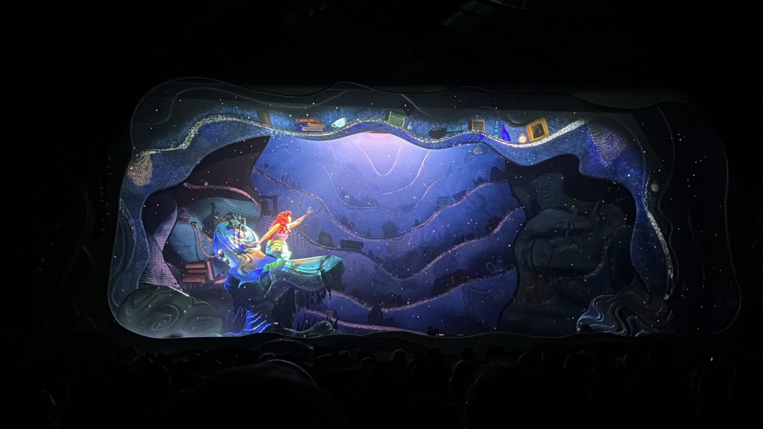 A theatrical stage displays an underwater scene from Little Mermaid: A Musical Adventure, with vibrant lighting and ocean-themed decor.