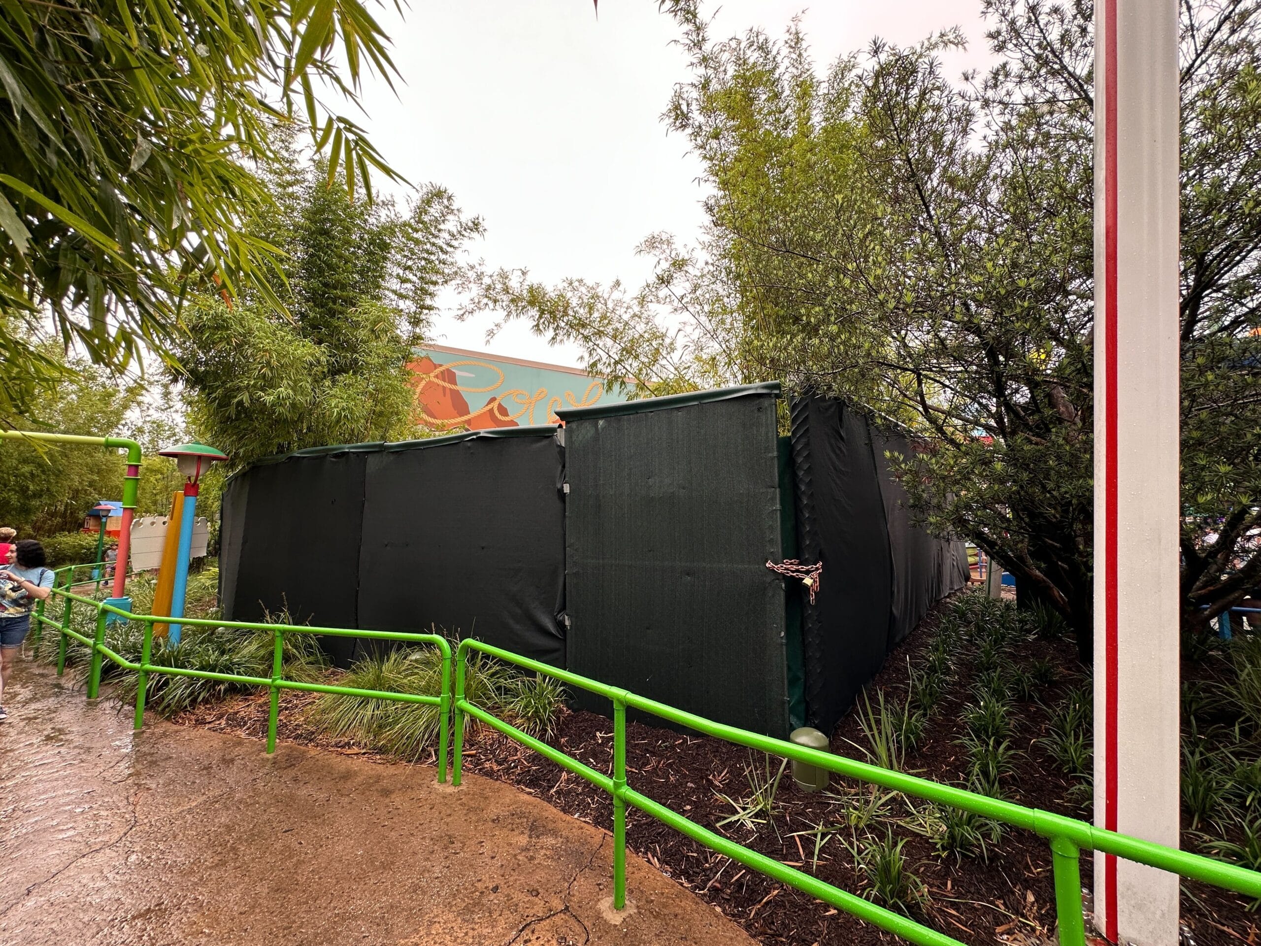 Green construction walls block off an area near plants and a sign, with a rainy path and green railing in front.