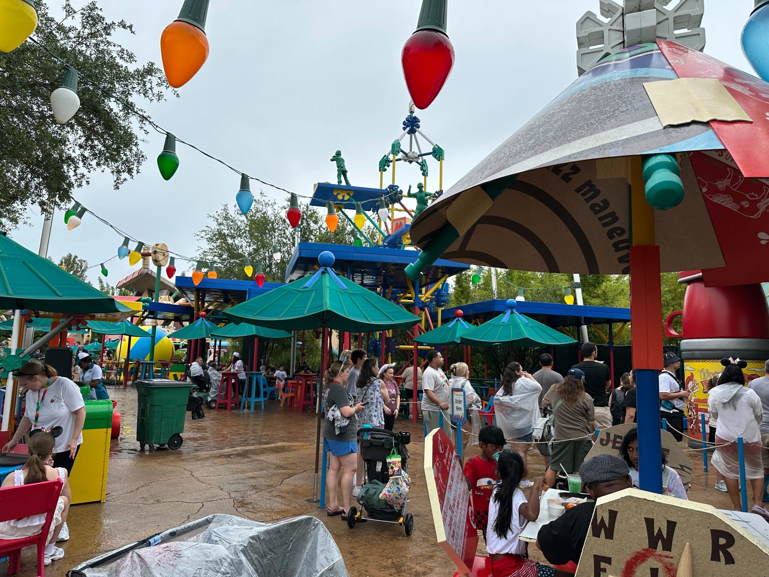 People walk and sit under umbrellas in a colorful, toy-themed outdoor area decorated with giant string lights.