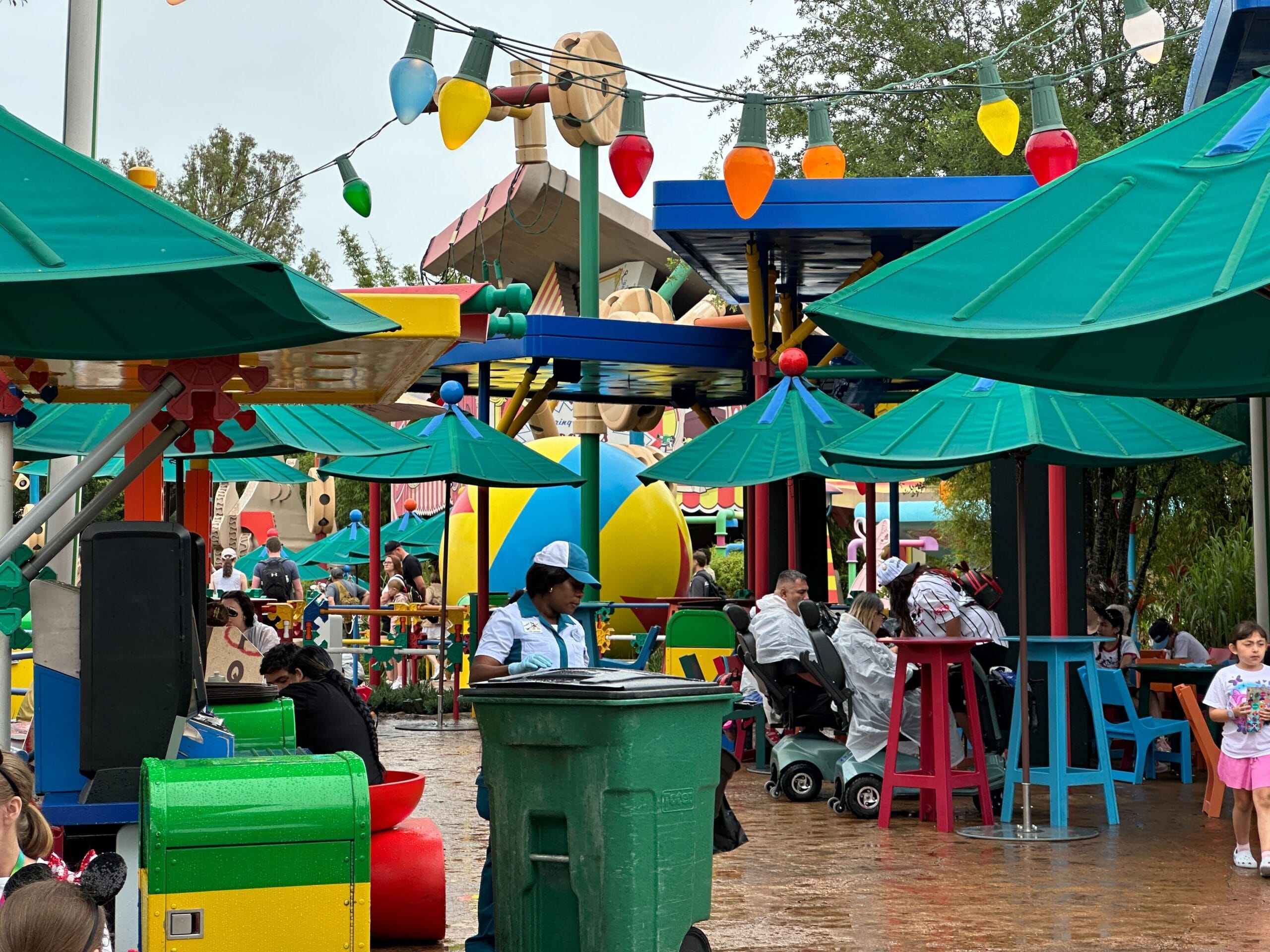Colorful outdoor seating area with green umbrellas, people, and large toy-themed decorations in a theme park.