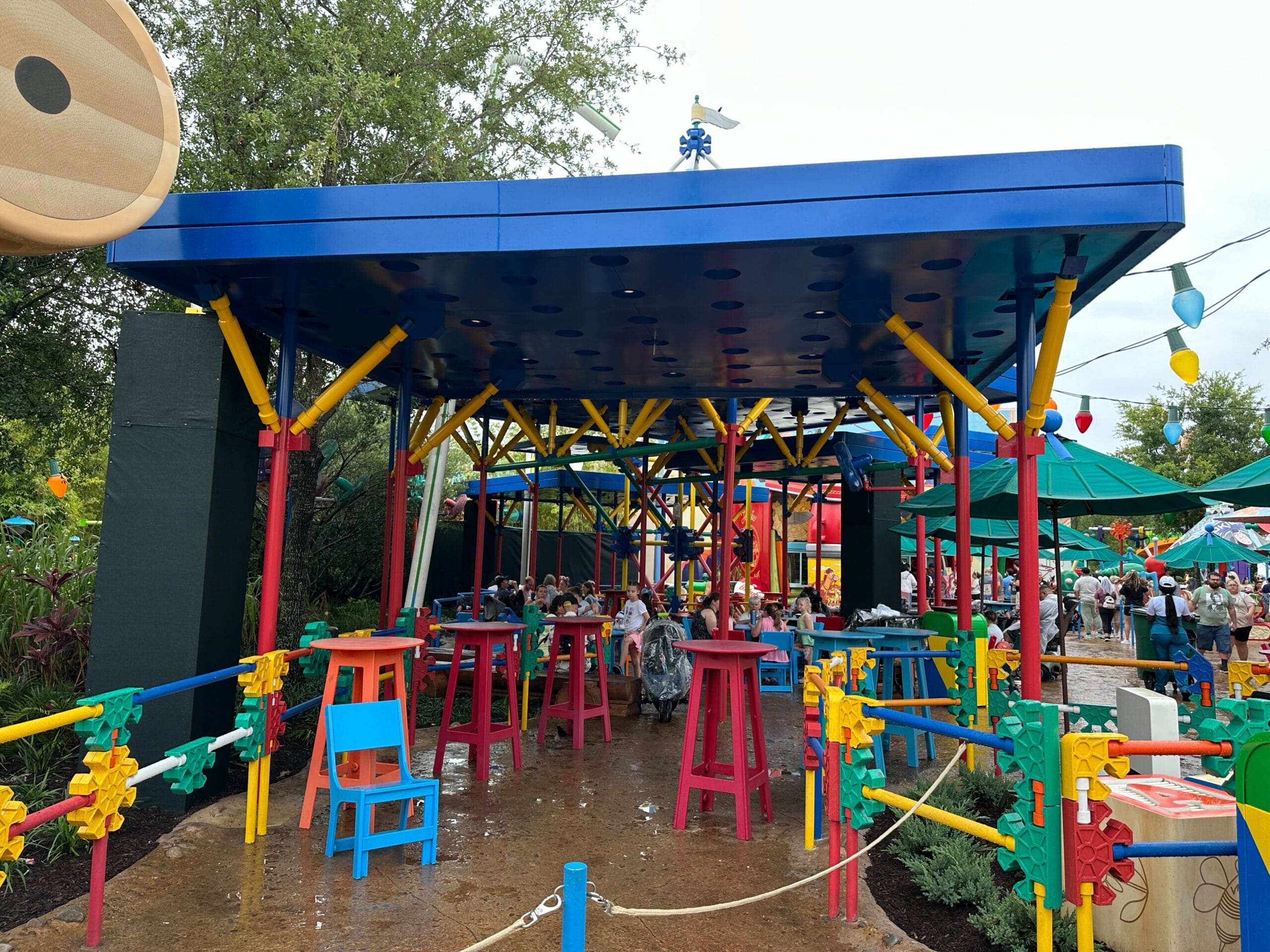Outdoor seating area with colorful tables, chairs, and toy-inspired structures, surrounded by greenery and playful decor.