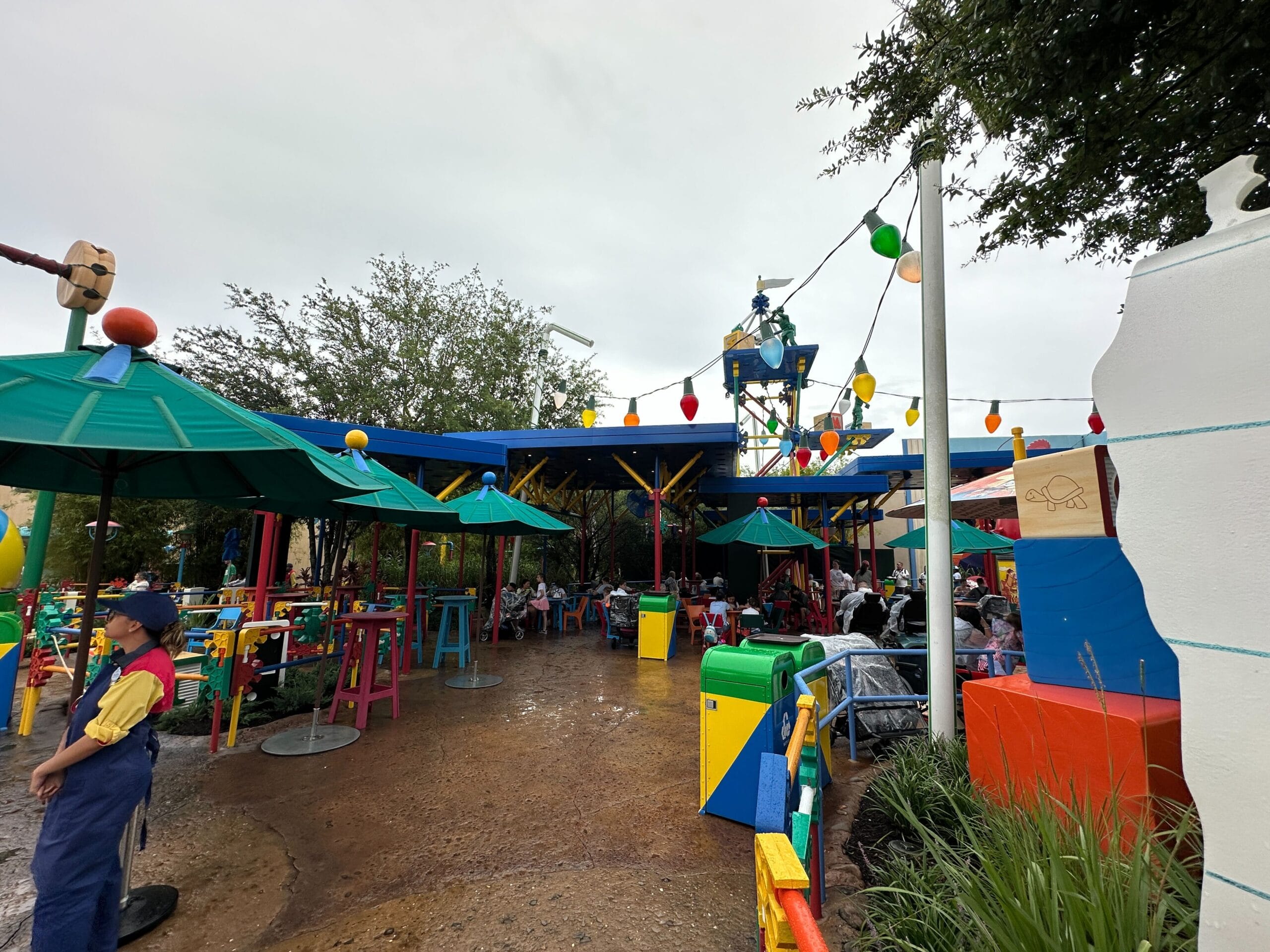 Outdoor dining area with colorful tables, umbrellas, string lights, and people sitting under a cloudy sky.