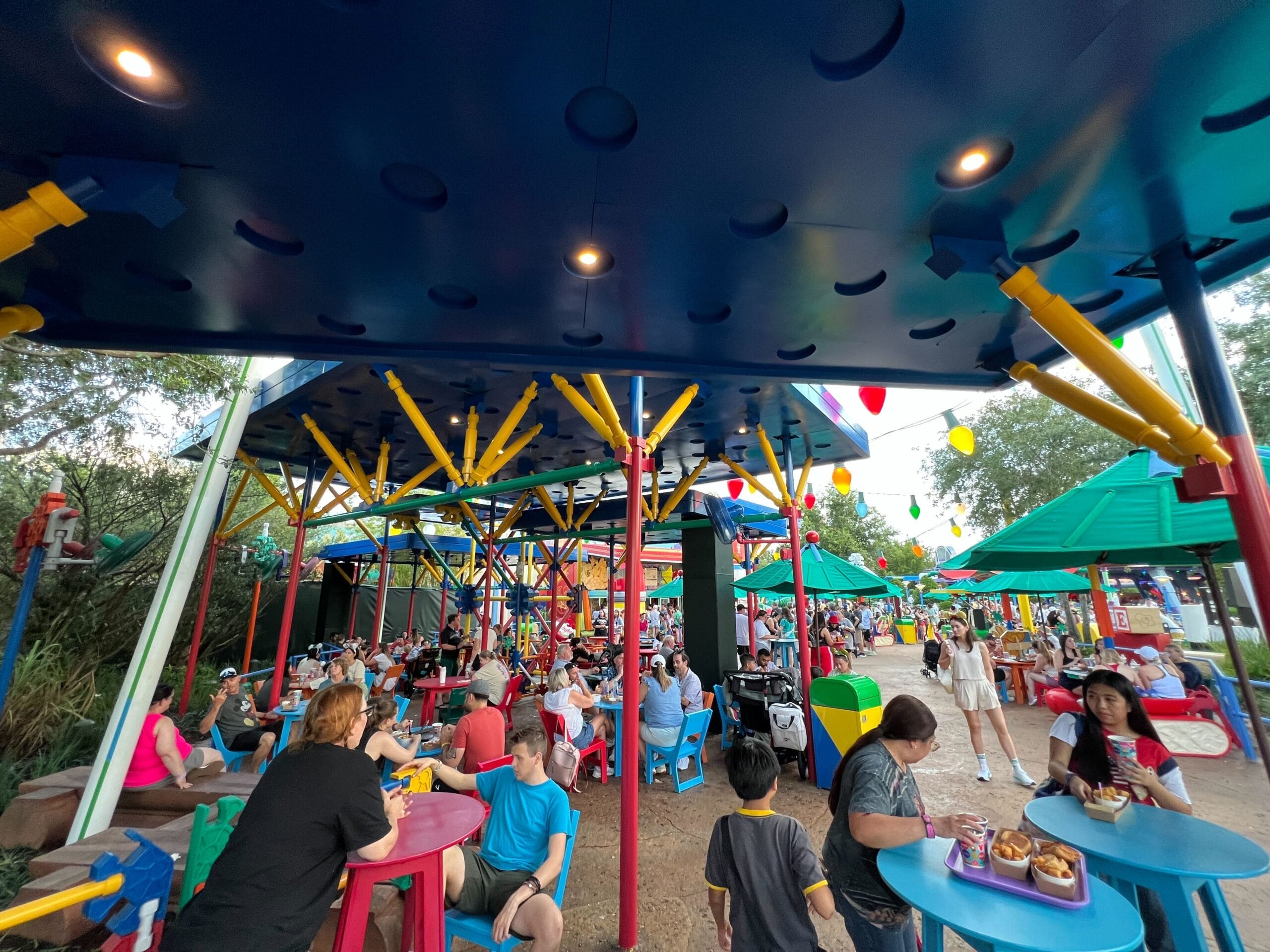 People dining outdoors at colorful tables under a blue canopy decorated with playful, oversized toy-themed structures.