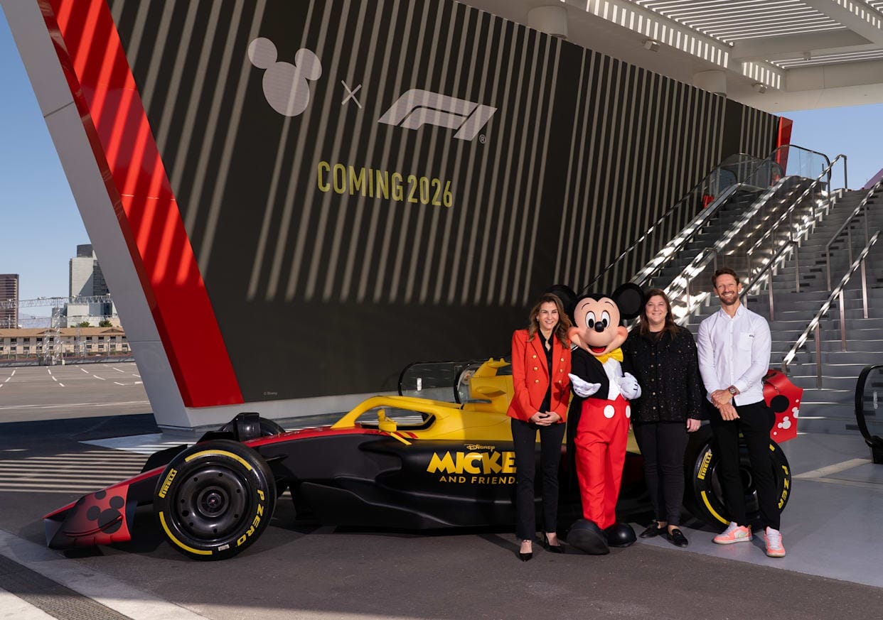 Four people, including Mickey Mouse, stand by a Disney-themed Formula 1 car; sign reads "Coming 2026.